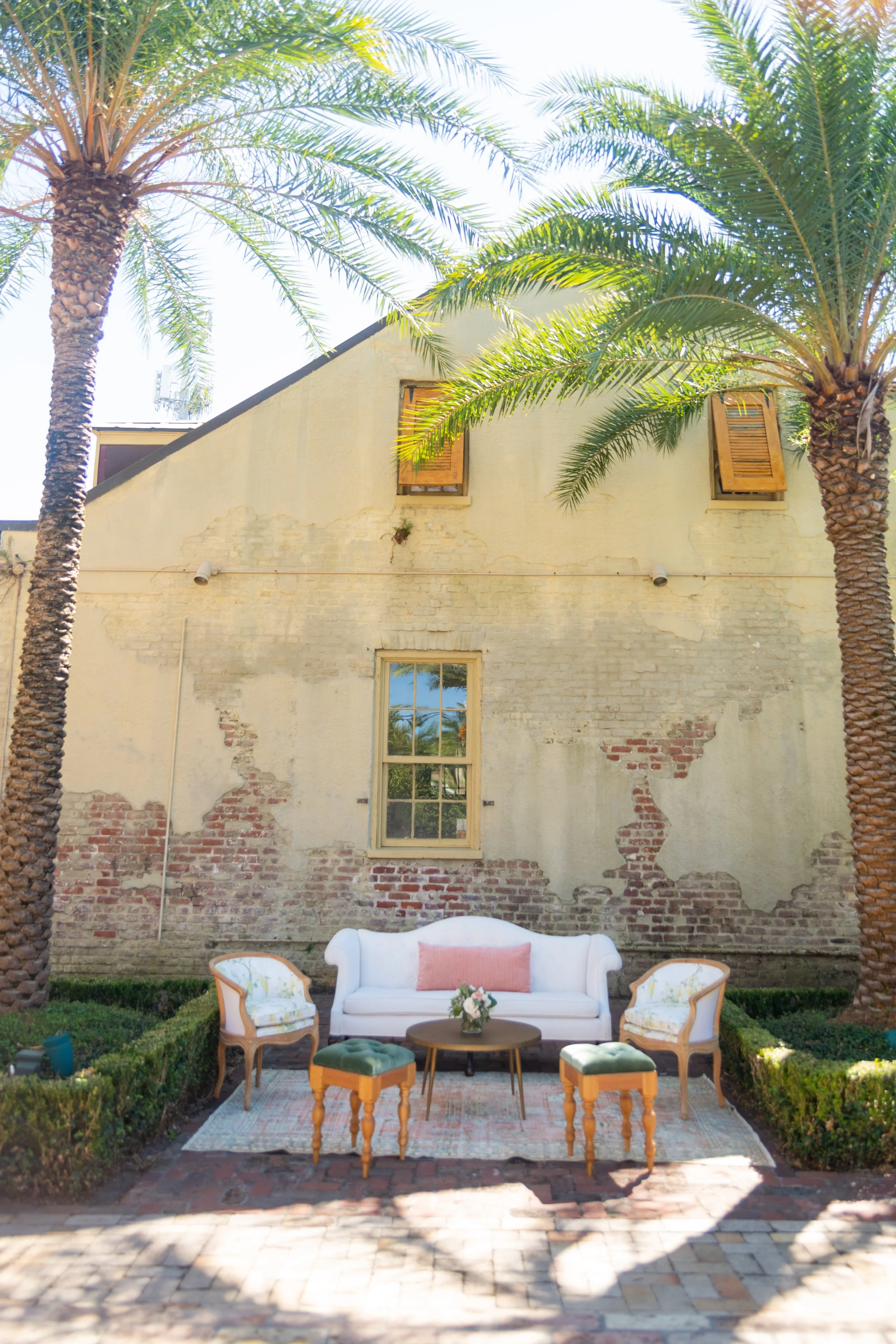 Outdoor patio setup with a white sofa, two armchairs, and a small round table with a flower arrangement, surrounded by green hedges and two tall palm trees, against a weathered brick wall and yellow building with open wooden window shutters.