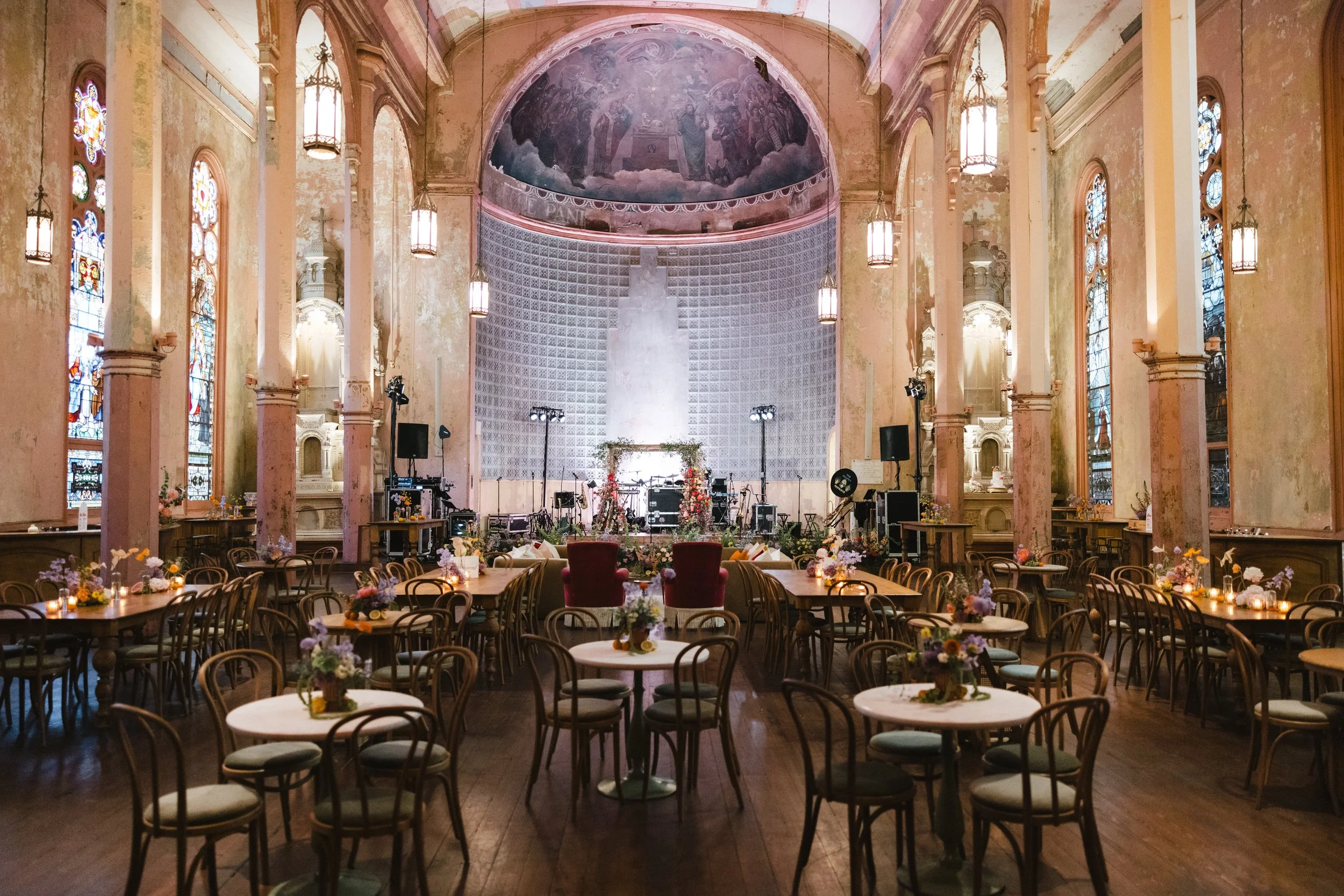 Interior of a church or cathedral with high, arched ceilings, stained glass windows, and hanging lantern lights. The space is set up for a wedding or event with tables, chairs, and floral arrangements, and a stage at the front.
