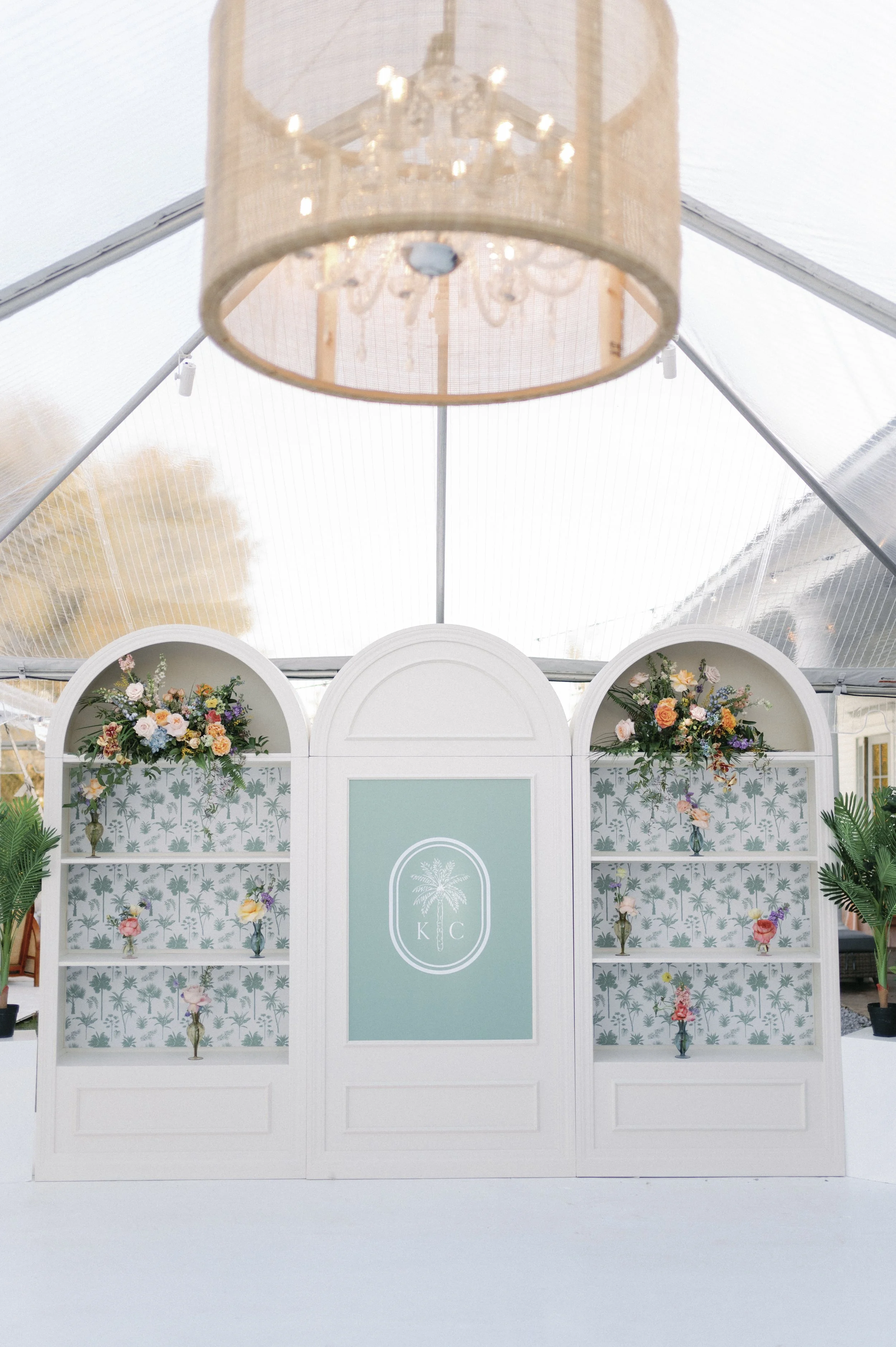 Decorative display with three white shelves filled with colorful flower arrangements, a mint green panel with palm tree graphic and initials 'K C' in a white frame, set inside a white room with a chandelier hanging from above.