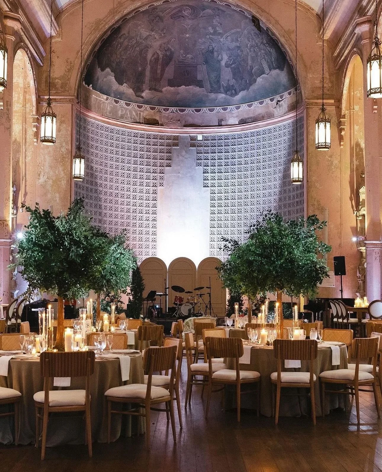 Elegant banquet hall decorated with green topiary trees, lit candles, and set tables with white tablecloths and chairs. The stage at the front has musical instruments, and a large mural is painted on the curved wall above the stage.