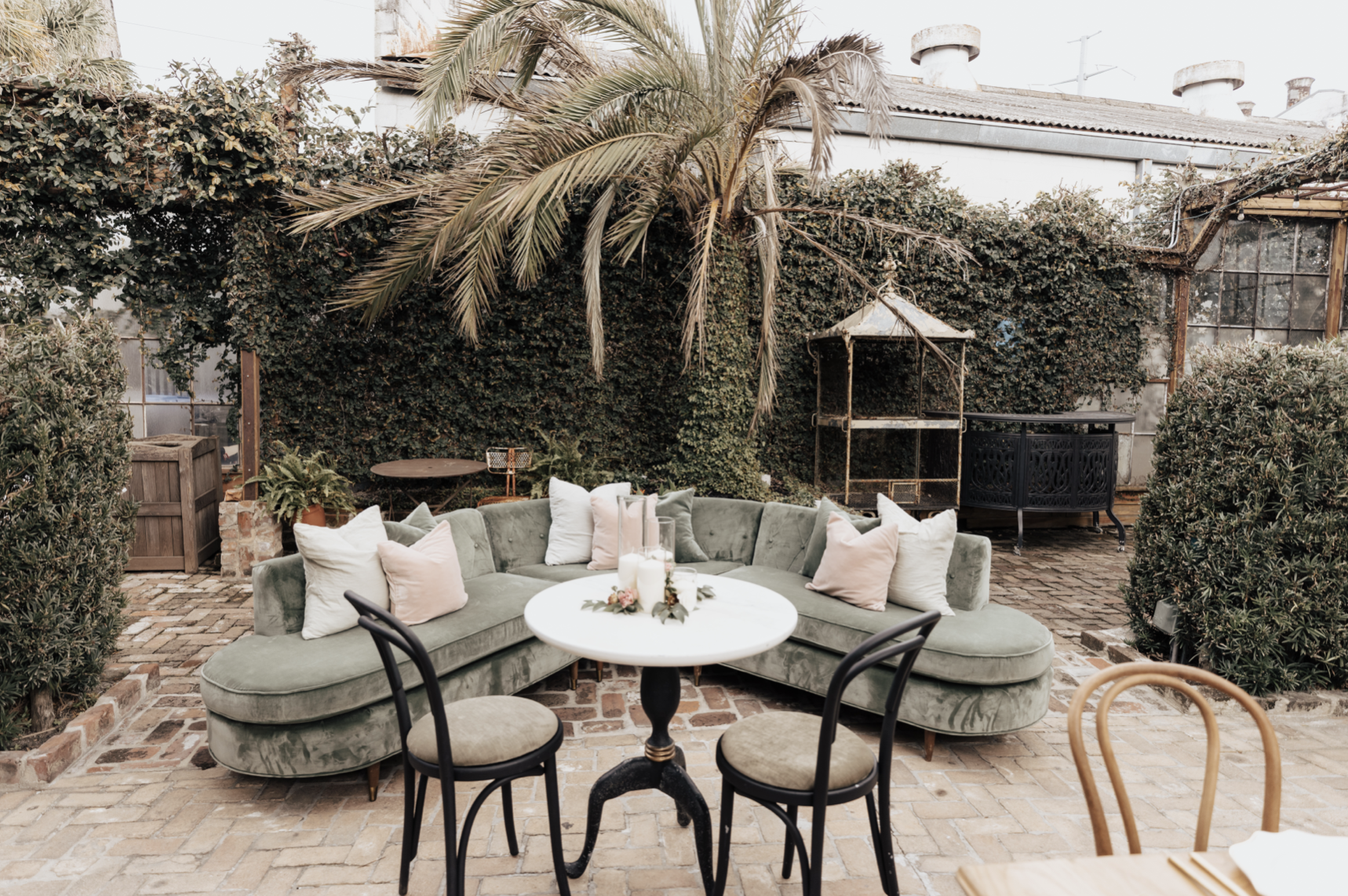 Outdoor patio with a green velvet sectional sofa adorned with white and pink pillows, a round white table with candles and greenery decor, surrounded by black and wooden chairs, brick paving, lush green plants, ivy-covered wall, and a small metal gaz