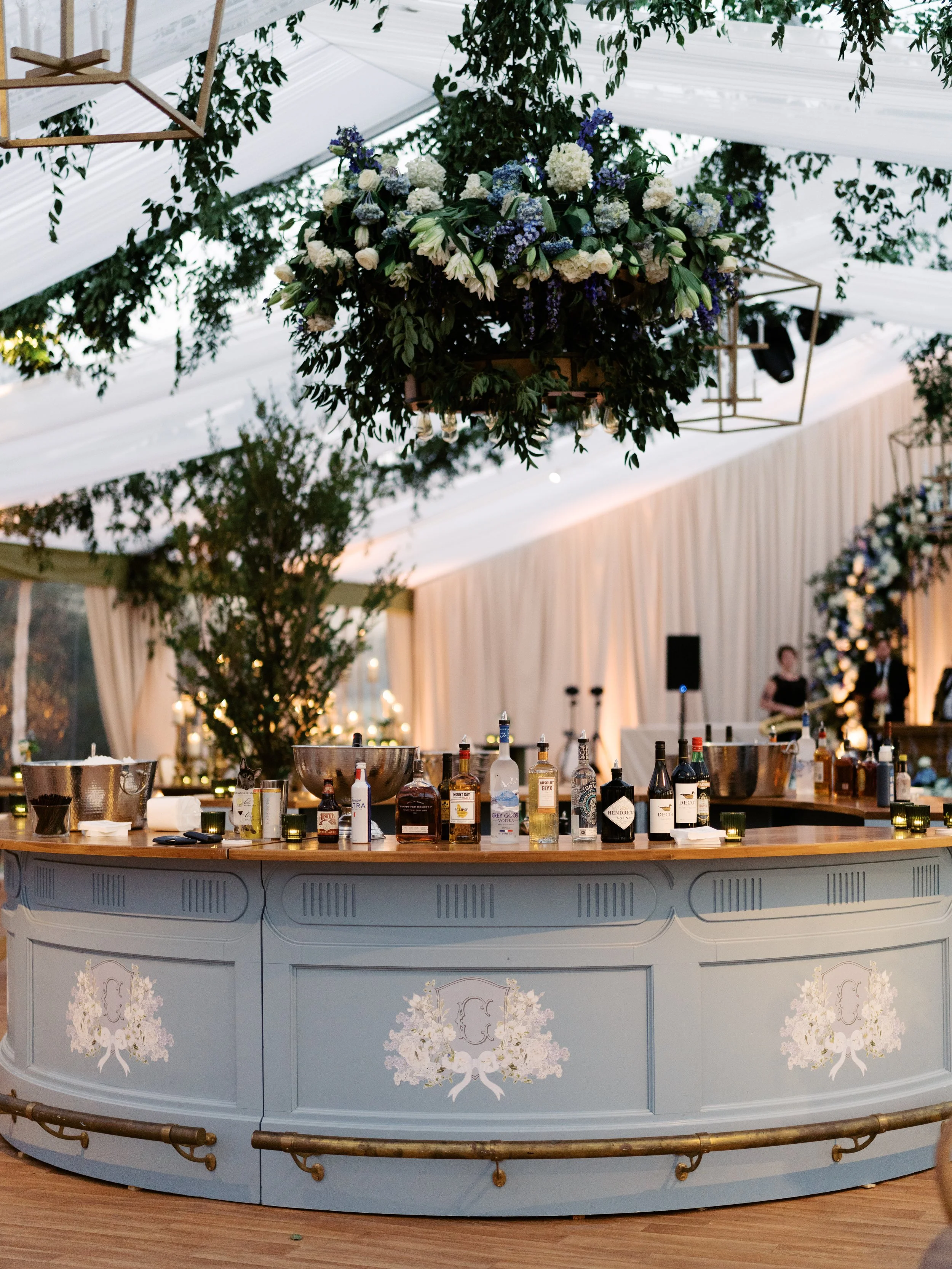 A round bar with various bottles of liquor on top, decorated with floral patterns and white floral designs on the front panel, located inside a tent with hanging flower arrangements and curtains.
