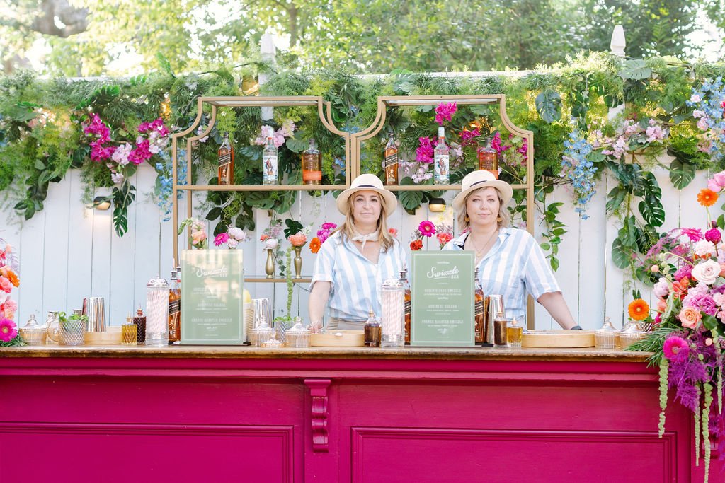 Two women standing behind a bar decorated with pink and purple flowers, greenery, and gold accents, under a canopy of trees.