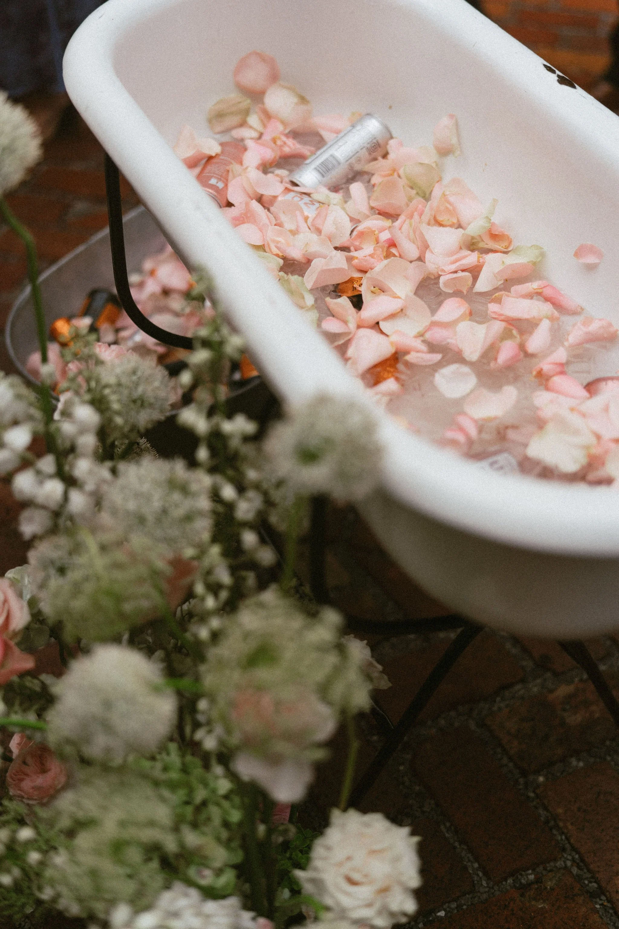 A white basin containing pink and white flower petals and a small aluminum can, placed on a table with a brick surface, surrounded by white flowers.