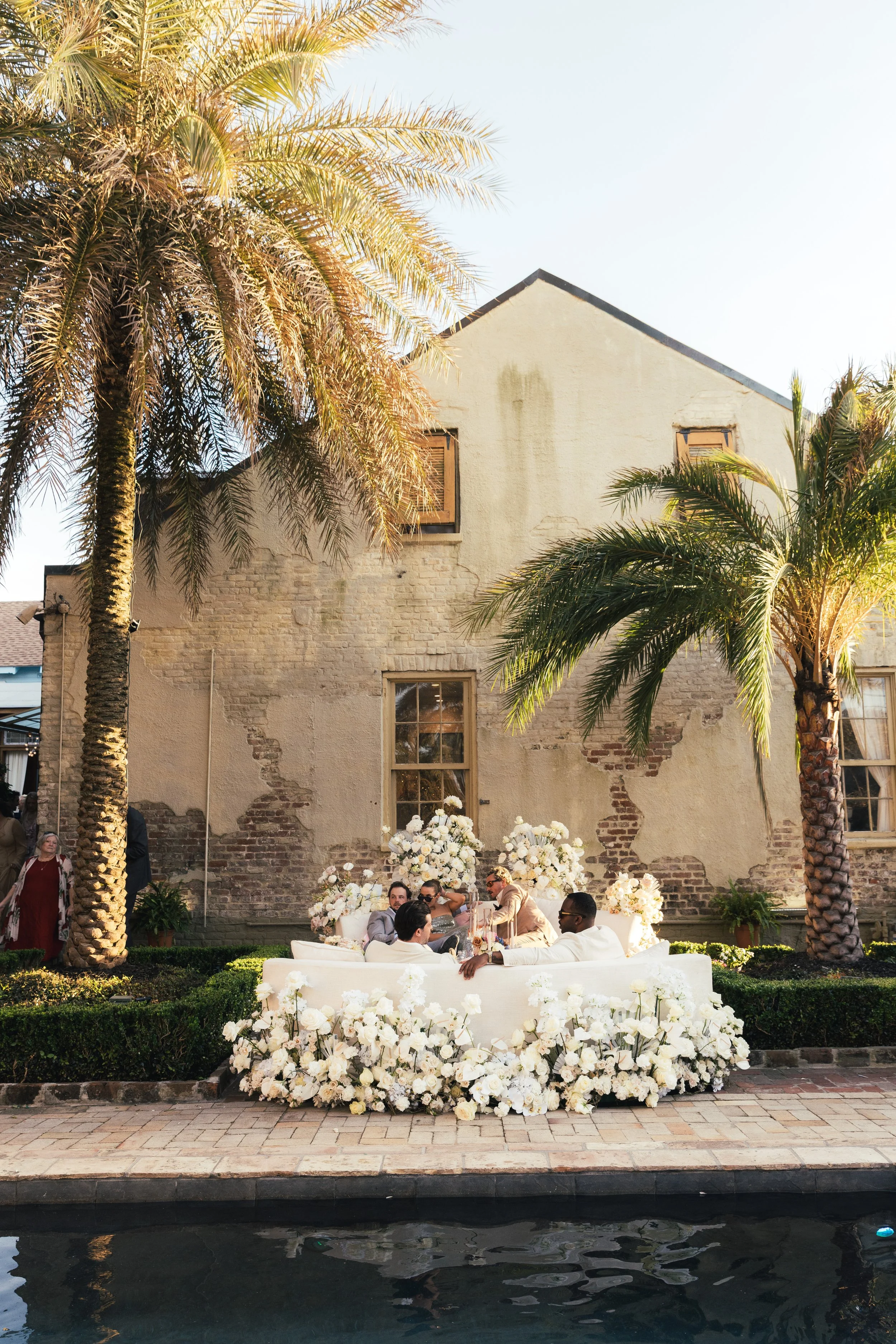 A group of people at a celebration or wedding reception outdoors, sitting around a table decorated with white flowers, with large palm trees and an old brick building in the background and a waterway at the bottom of the image.