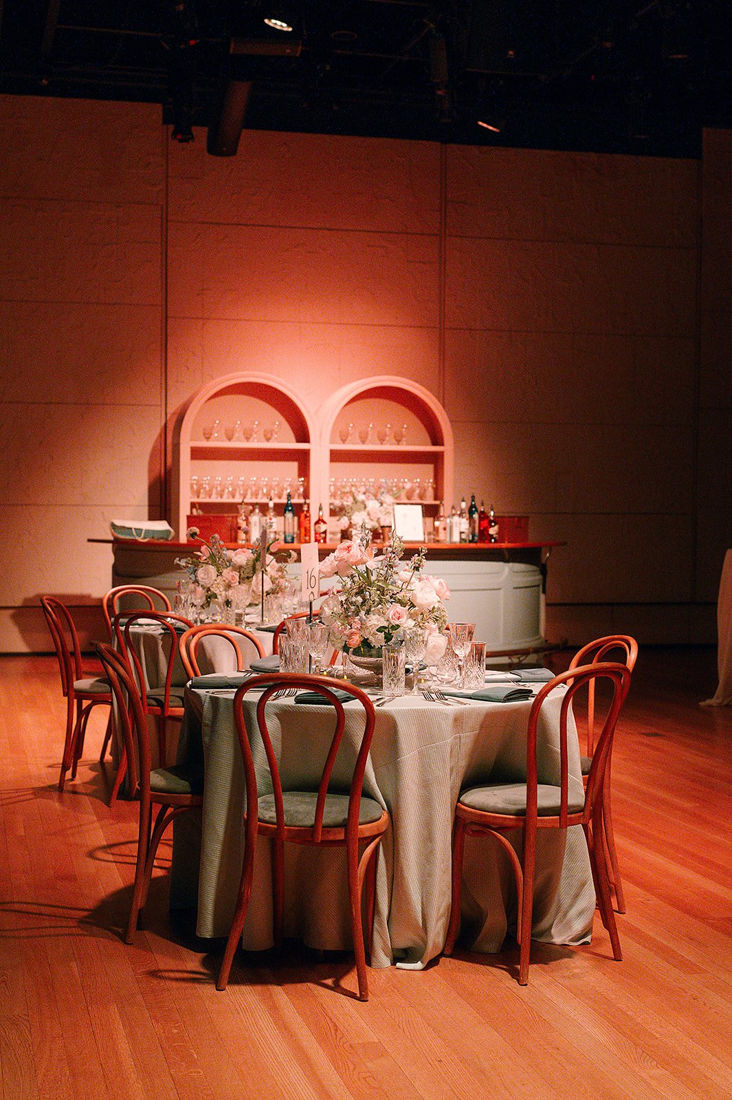 Elegant event setup with a round table decorated with pink and white flowers, surrounded by wooden chairs with light-colored cushions, on a wooden floor. In the background, a bar with bottles and glassware is set against a pink wall with arched shelv