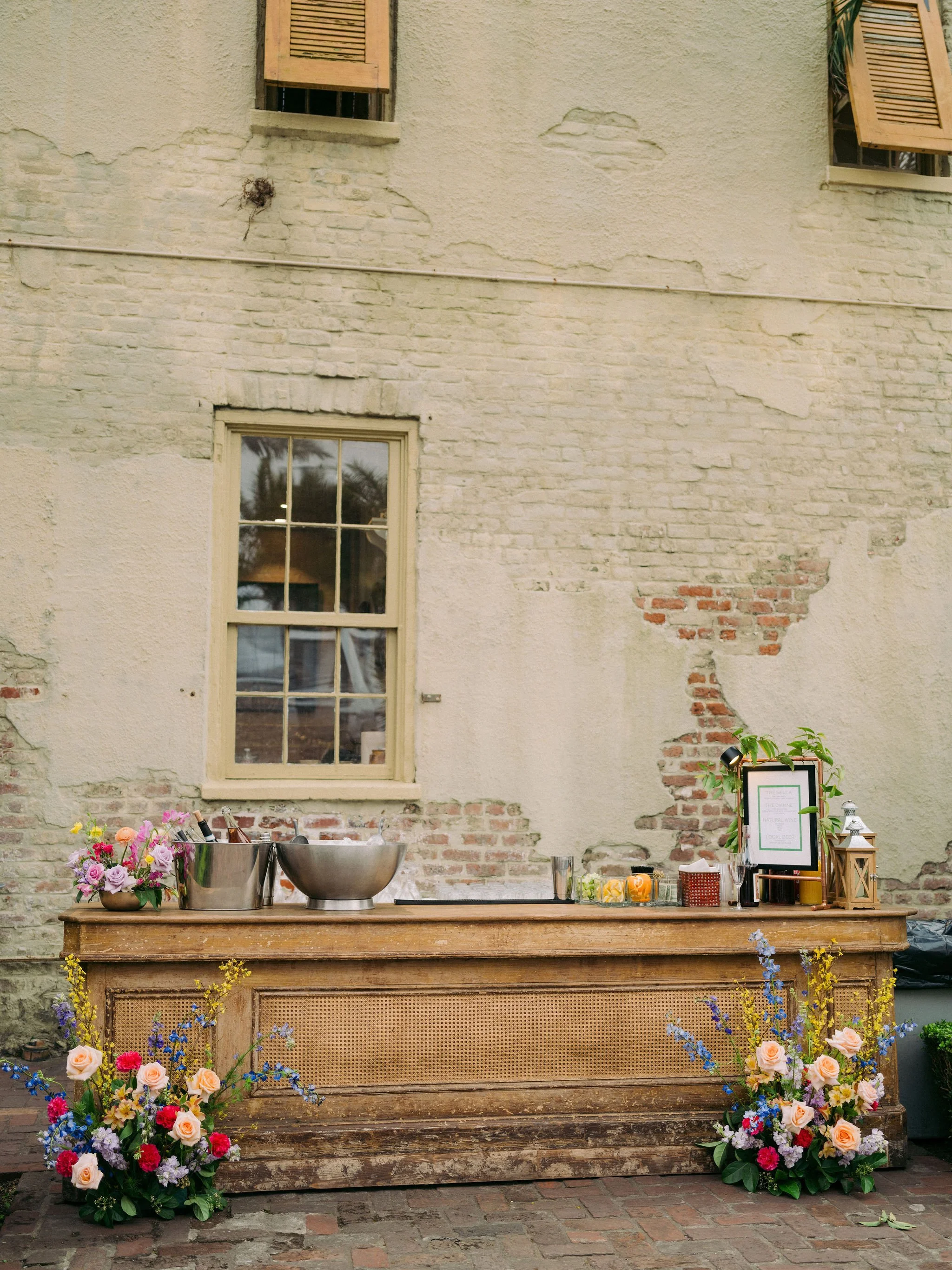 A rustic outdoor bar setup against a weathered brick and beige stucco wall with open window shutters. The wooden bar has floral arrangements, a metal ice bucket, citrus slices in cups, and signs and lanterns.