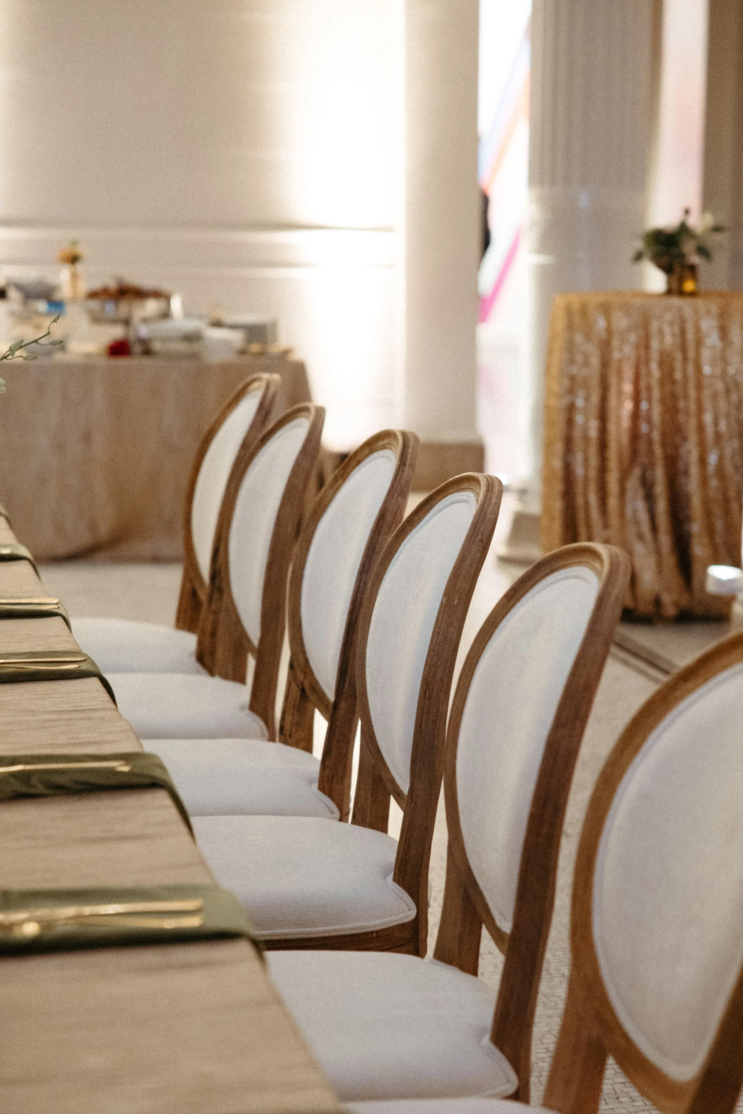 Elegant event space with a row of white cushioned chairs with wooden frames, a long table set with gold-edged napkins, and blurred background featuring a decorated table and floral arrangements. Bright light coming from windows or doors.