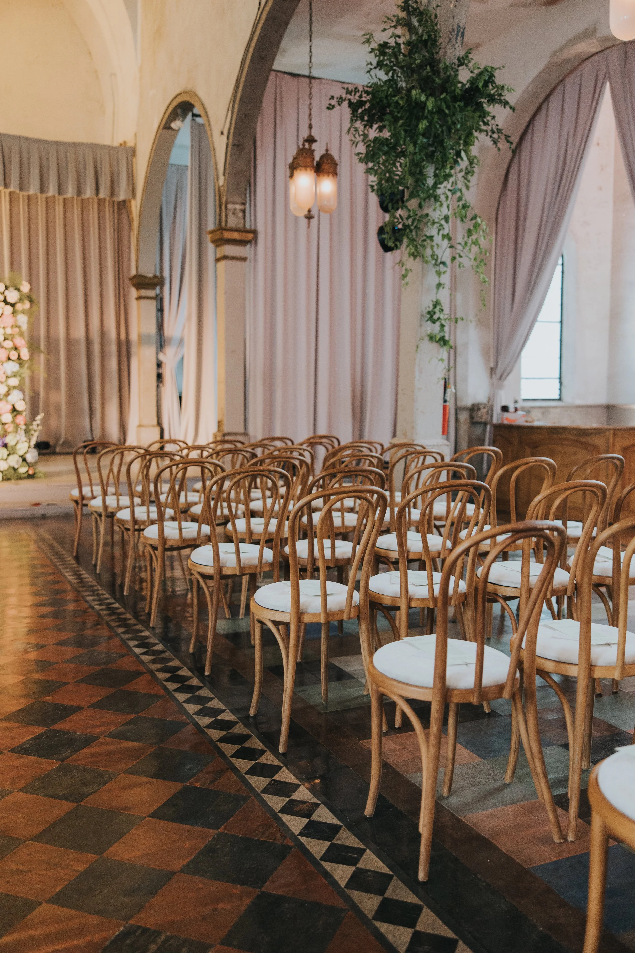 Interior of a decorated event space with rows of wooden chairs with white cushions, ornate curtains, large hanging lamps, a flower arrangement, and a green plant.