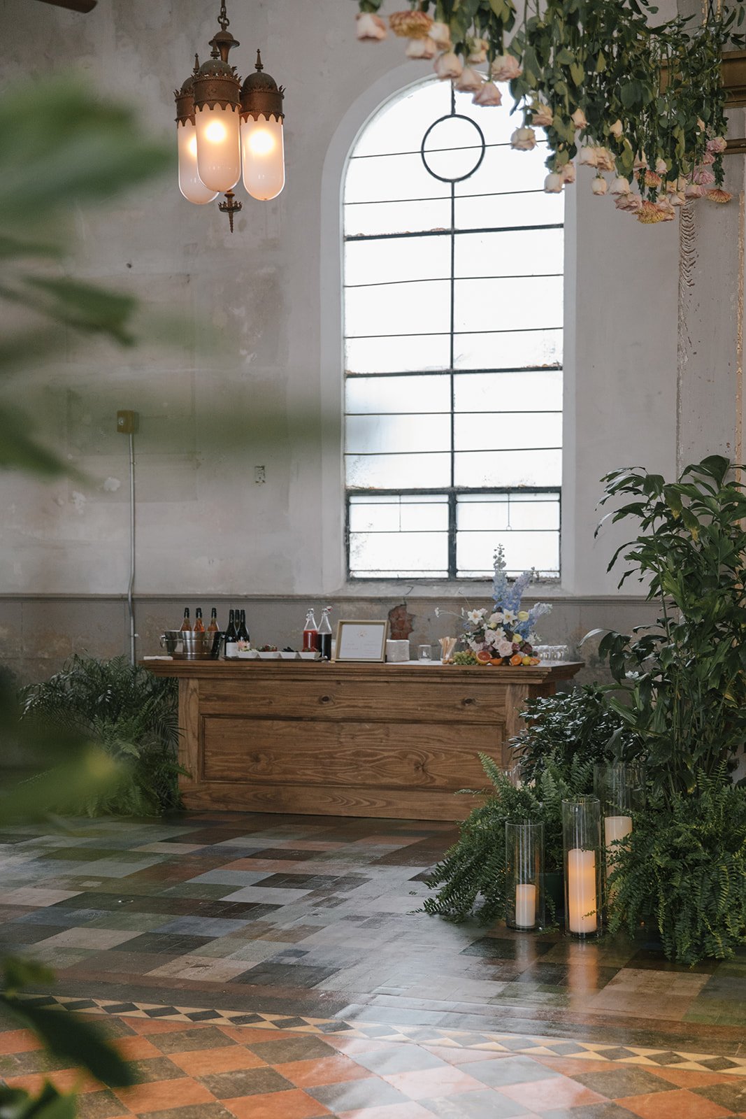 A rustic indoor space with a large arched window, hanging chandelier, green plants, candles, and a wooden table decorated with flowers, bottles, and framed items.