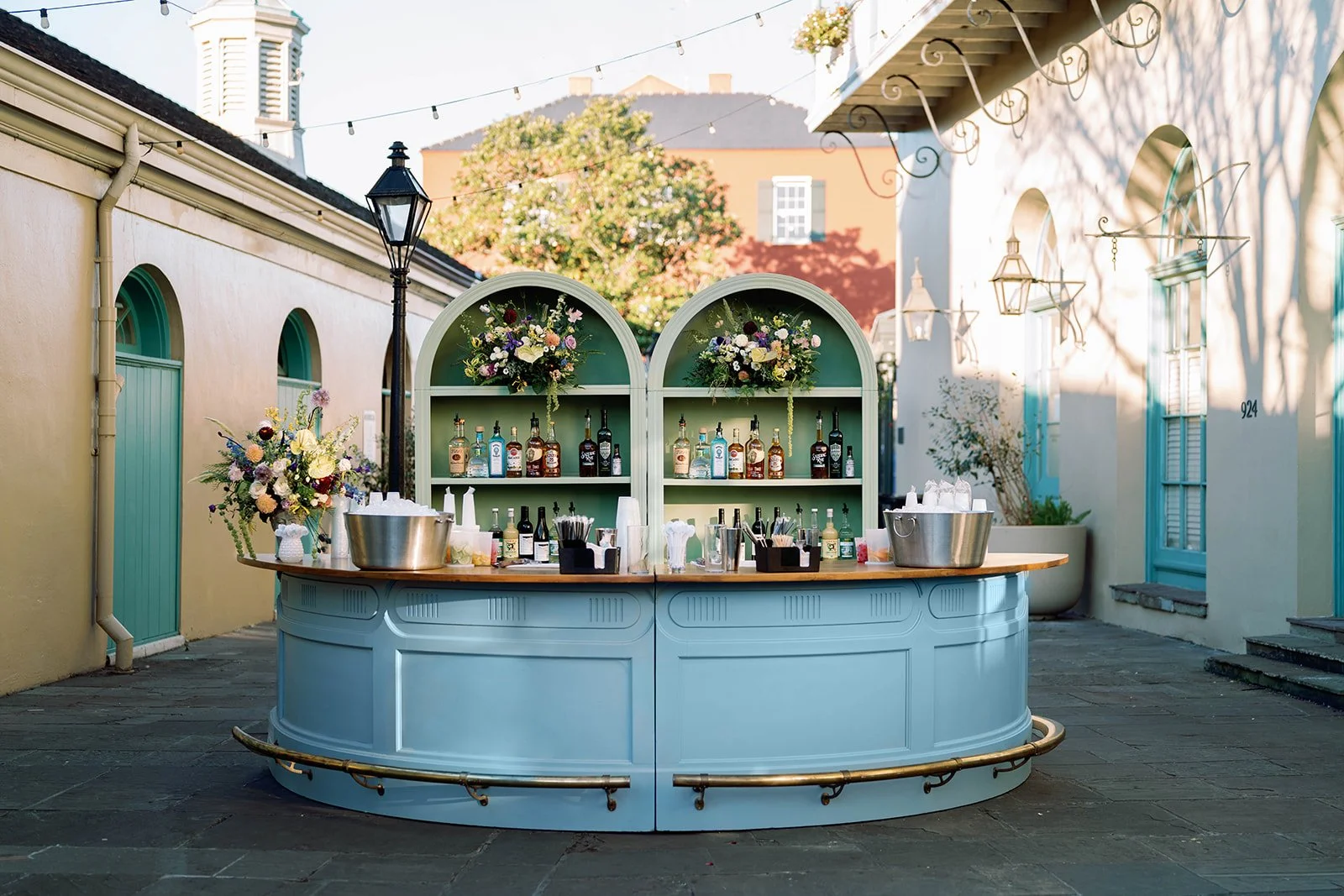 An outdoor bar with a light blue curved counter, bottles of alcohol on green shelves, floral arrangements in baskets, and large metal bowls on either side of the bar. String lights hang above, and there are pastel-colored buildings and trees in the b