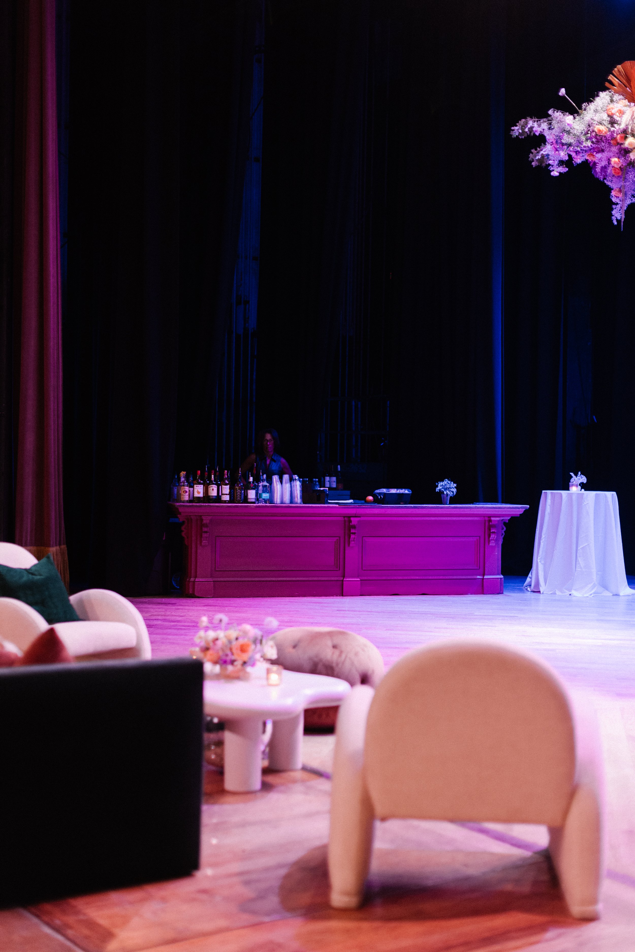 Interior of an event space with soft purple lighting, featuring a bar with bottles and drinks, a bartender, and a seating area with beige and dark upholstered chairs and tables with flower arrangements.