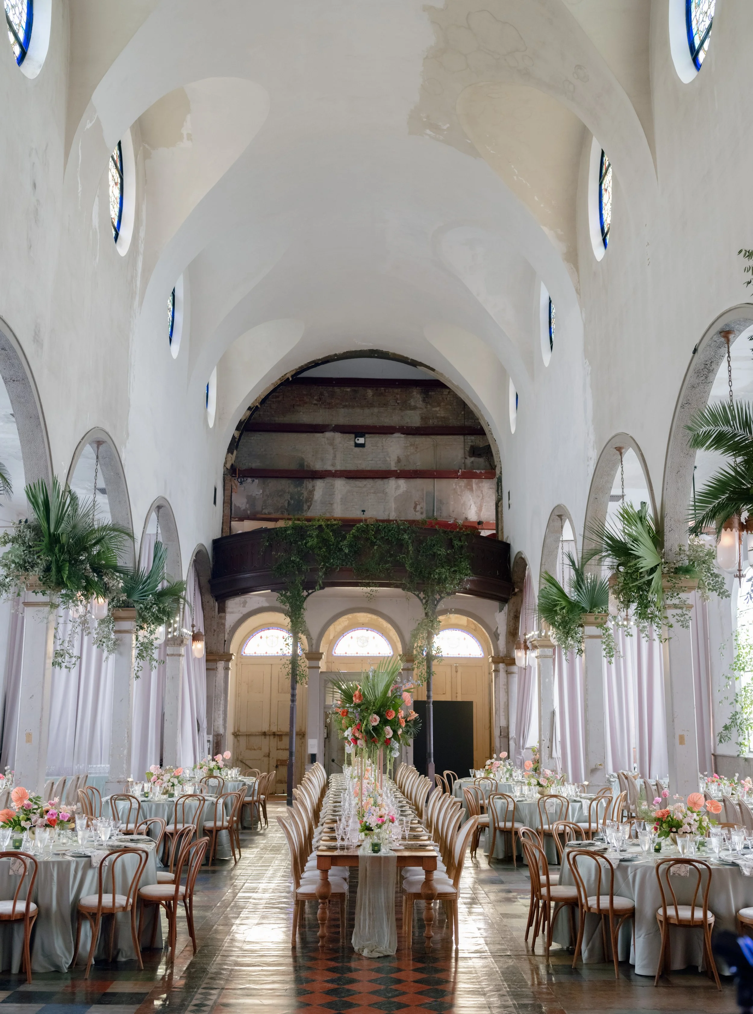 Elegant banquet hall decorated for a wedding with long tables, floral centerpieces, and high ceilings, inside a church with stained glass windows.