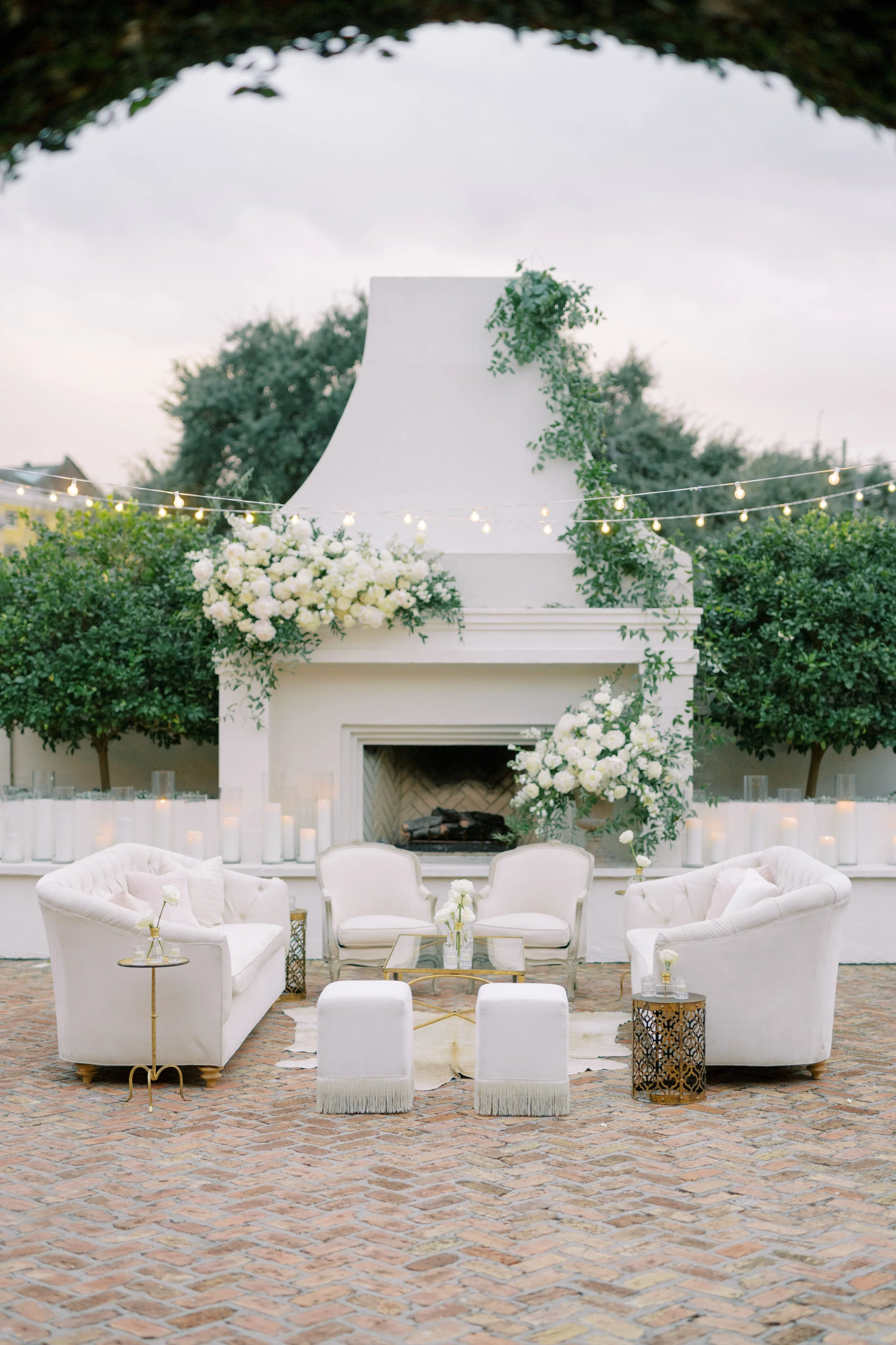 A outdoor wedding reception seating area with white sofas and armchairs, a glass coffee table, floral arrangements, small tables with candles, a fireplace decorated with flowers, string lights, greenery, and trees in the background at Il Mercato in N