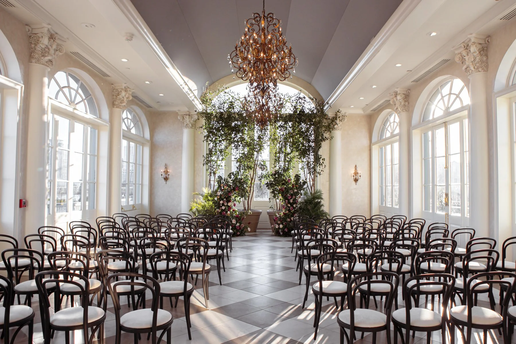 Elegant indoor wedding ceremony setup with rows of black chairs with white cushions facing an altar area decorated with flowers and greenery, large windows letting in natural light, a crystal chandelier hanging from the ceiling, and classic architect