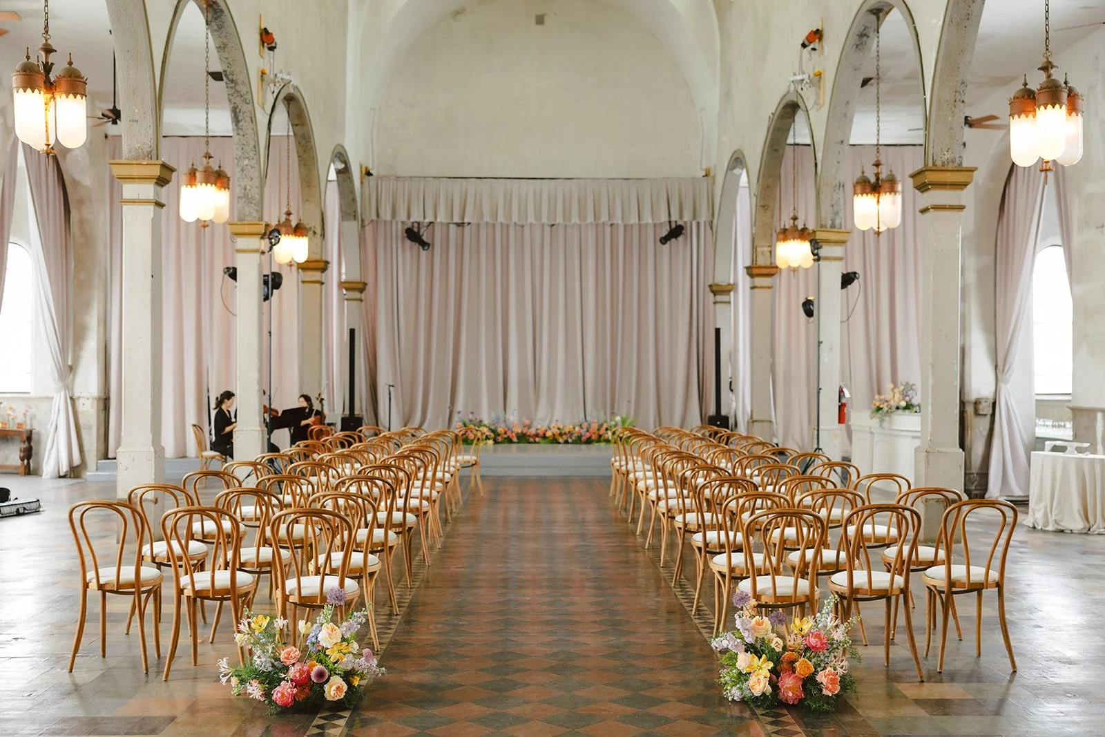Indoor wedding ceremony setup with rows of wooden chairs, flower arrangements, a stage with a curtain, and musicians preparing for the event.
