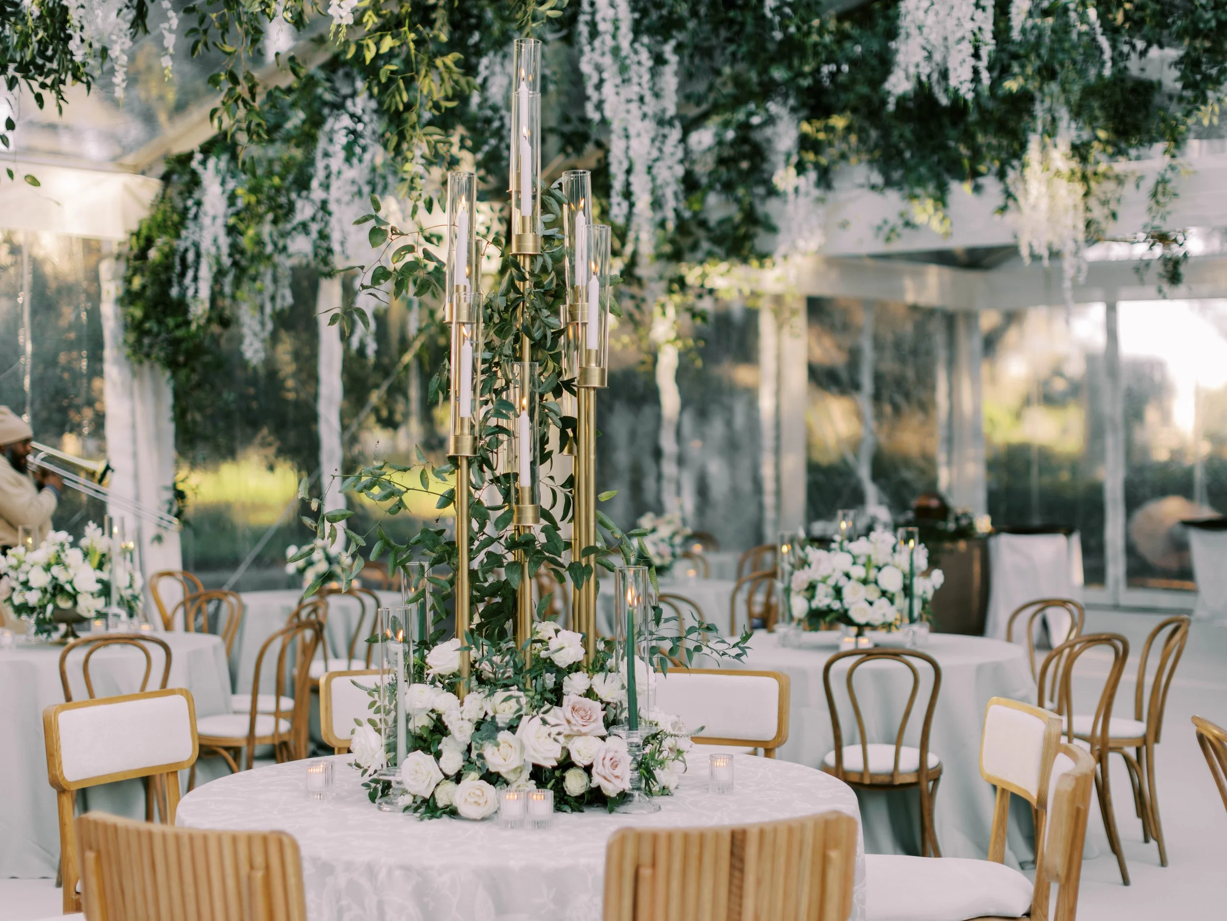 Elegant wedding reception setup with a round table decorated with white and pink flowers, candles, and a tall gold candelabra surrounded by greenery, under a glass ceiling with hanging white floral arrangements.