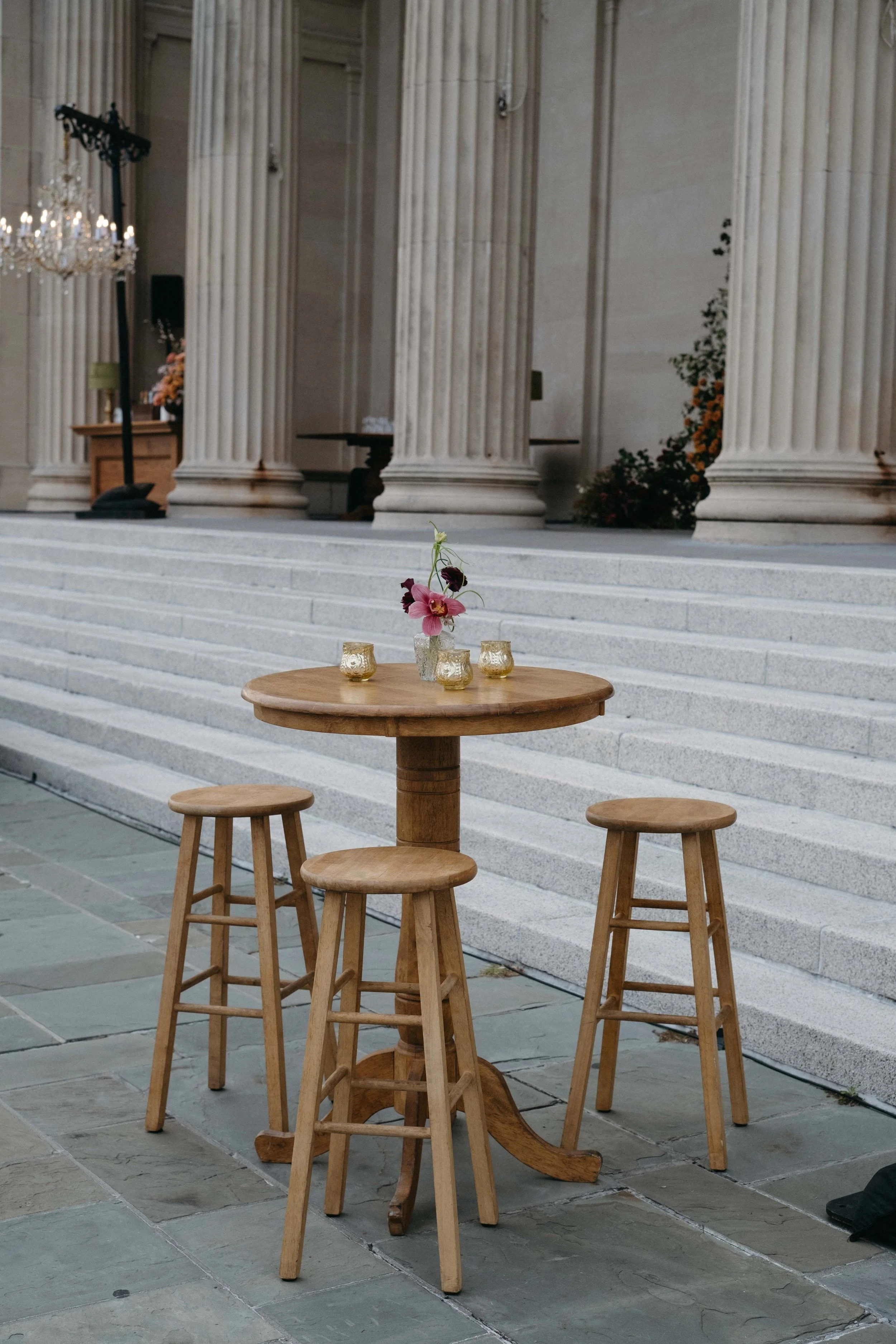 A round wooden table with four matching wooden stools placed on a stone-paved outdoor area in front of large stone steps leading up to a building with tall columns. The table has a small floral arrangement with pink flowers and four small candles in 