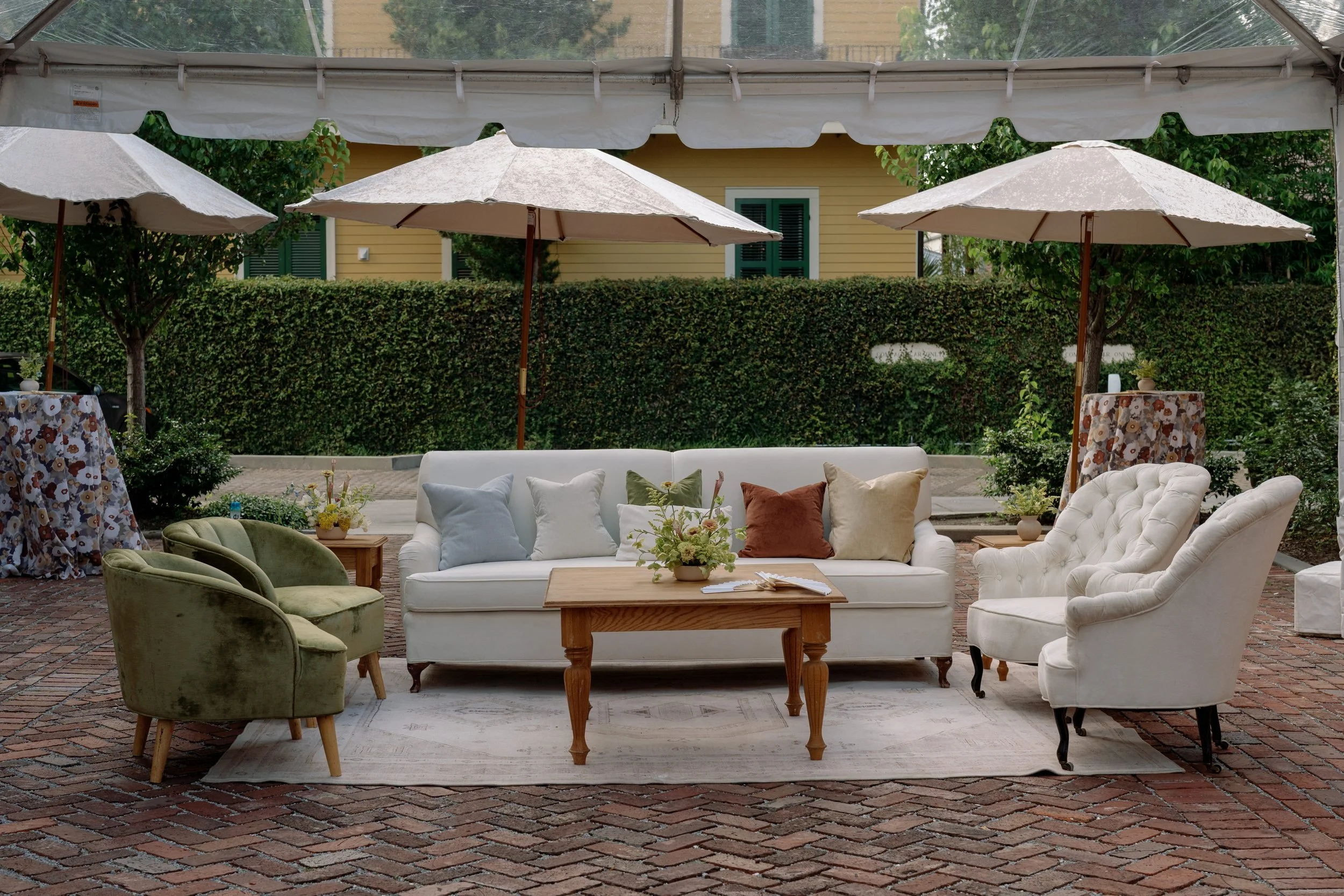 An outdoor patio setup with white, green, and beige chairs surrounding a wooden coffee table with a flower arrangement, beneath three large beige umbrellas, with a yellow house and green hedges in the background.