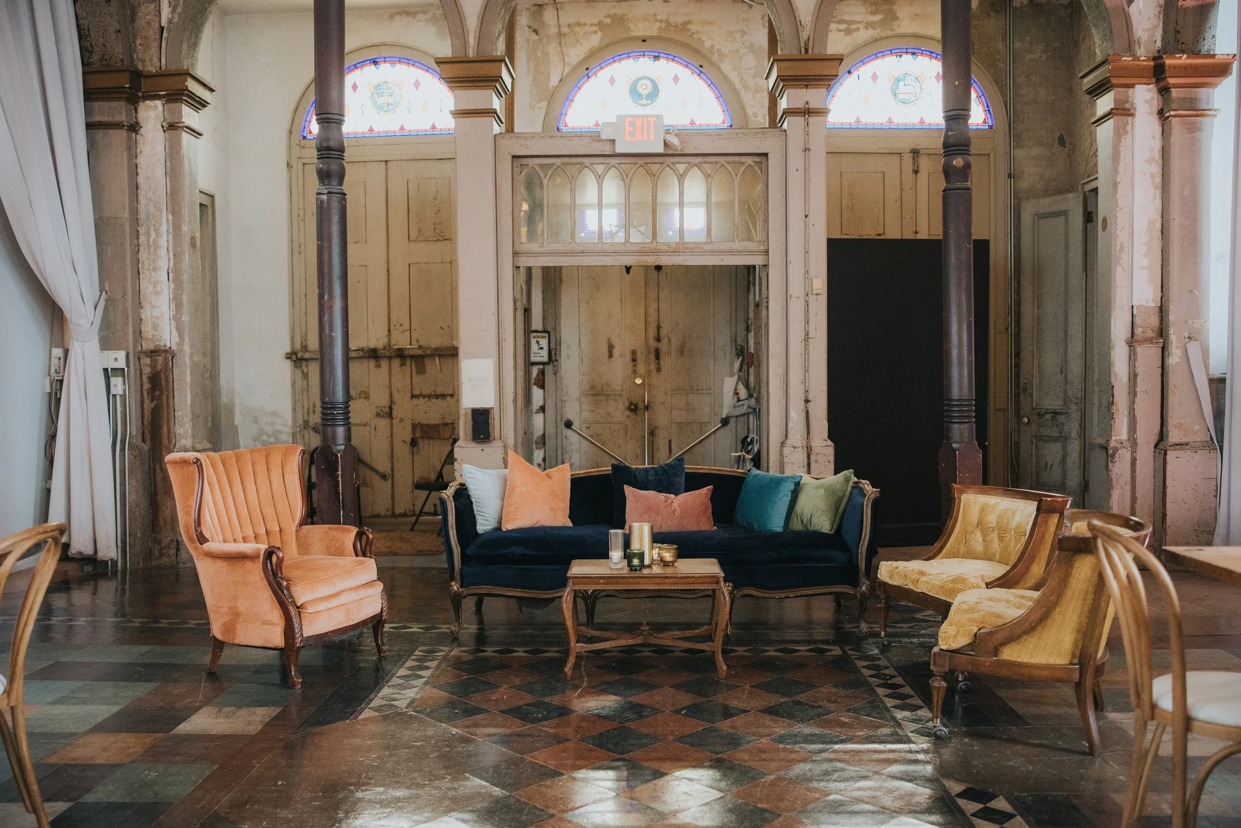 Interior of an old building with vintage furniture, including upholstered armchairs, a sofa with colorful pillows, and a coffee table with candles. The walls are worn and peeling, and stained glass windows are above a doorway marked with an exit sign