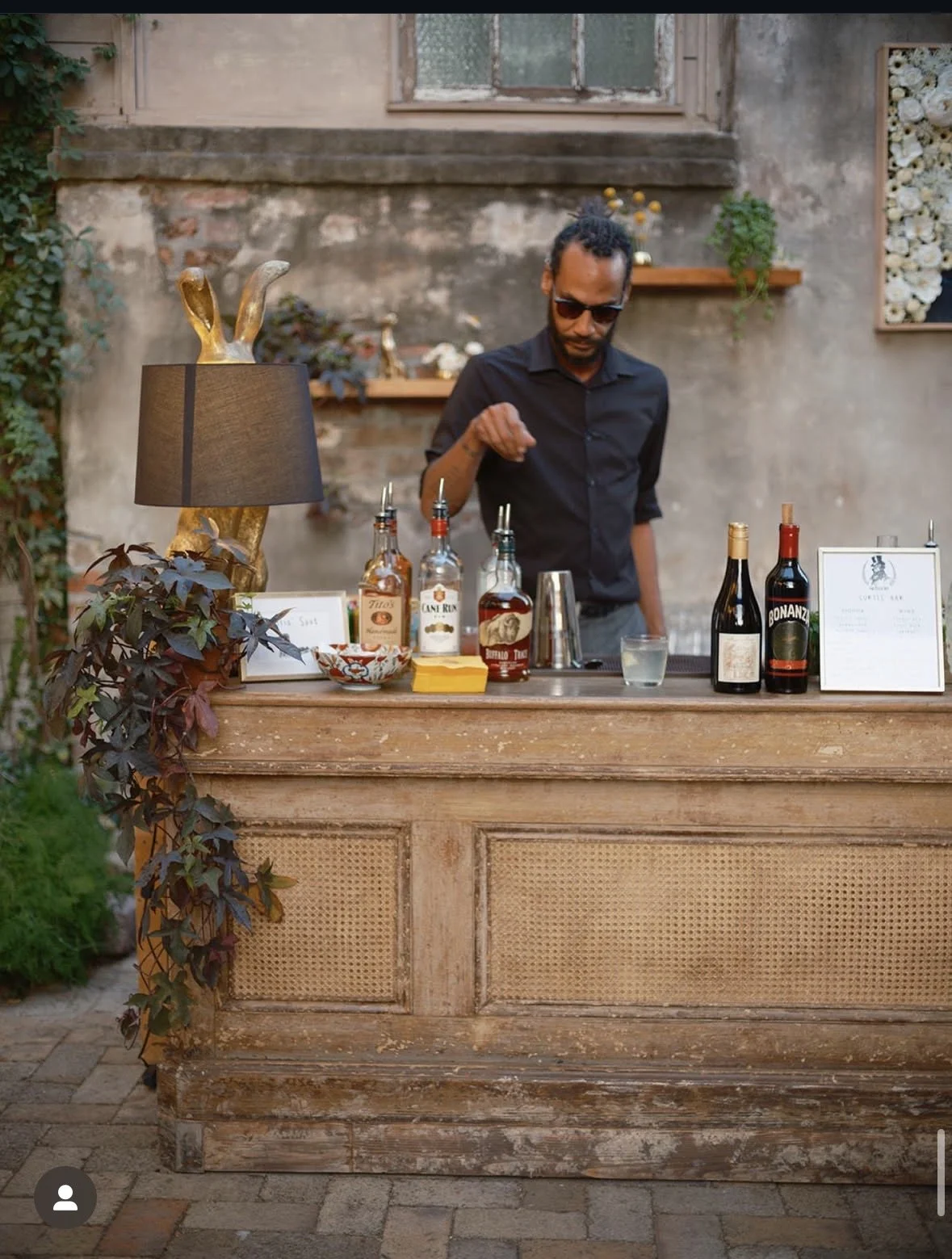 A bartender standing behind an outdoor bar setup with bottles of alcohol, a glass of water, and a cocktail shaker, in a rustic setting with a weathered wall and shelves with decorations.
