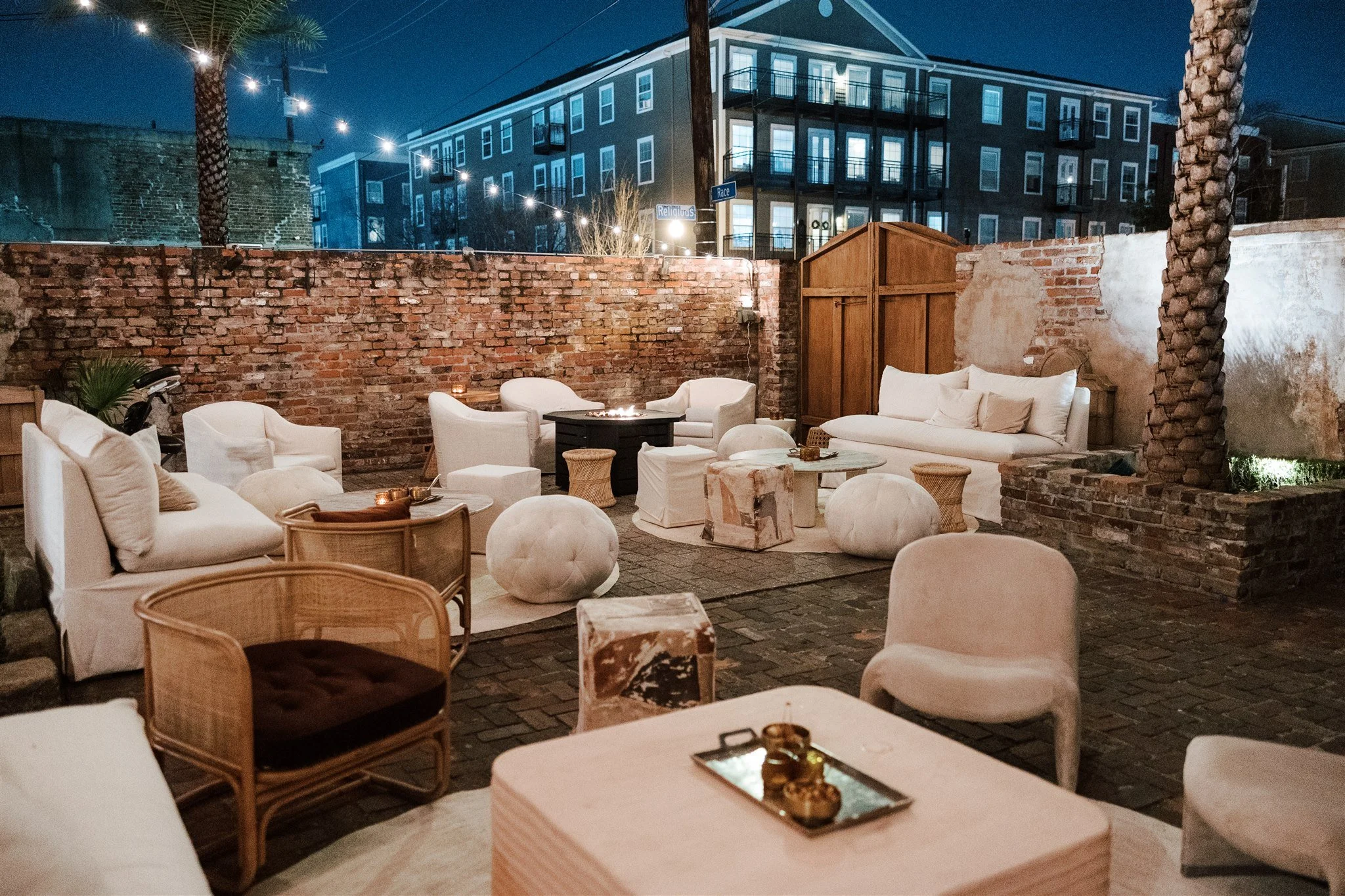 An outdoor seating area at night with white cushioned chairs and a sofa, a central fire pit, a brick wall, string lights, and a building with lit windows in the background.
