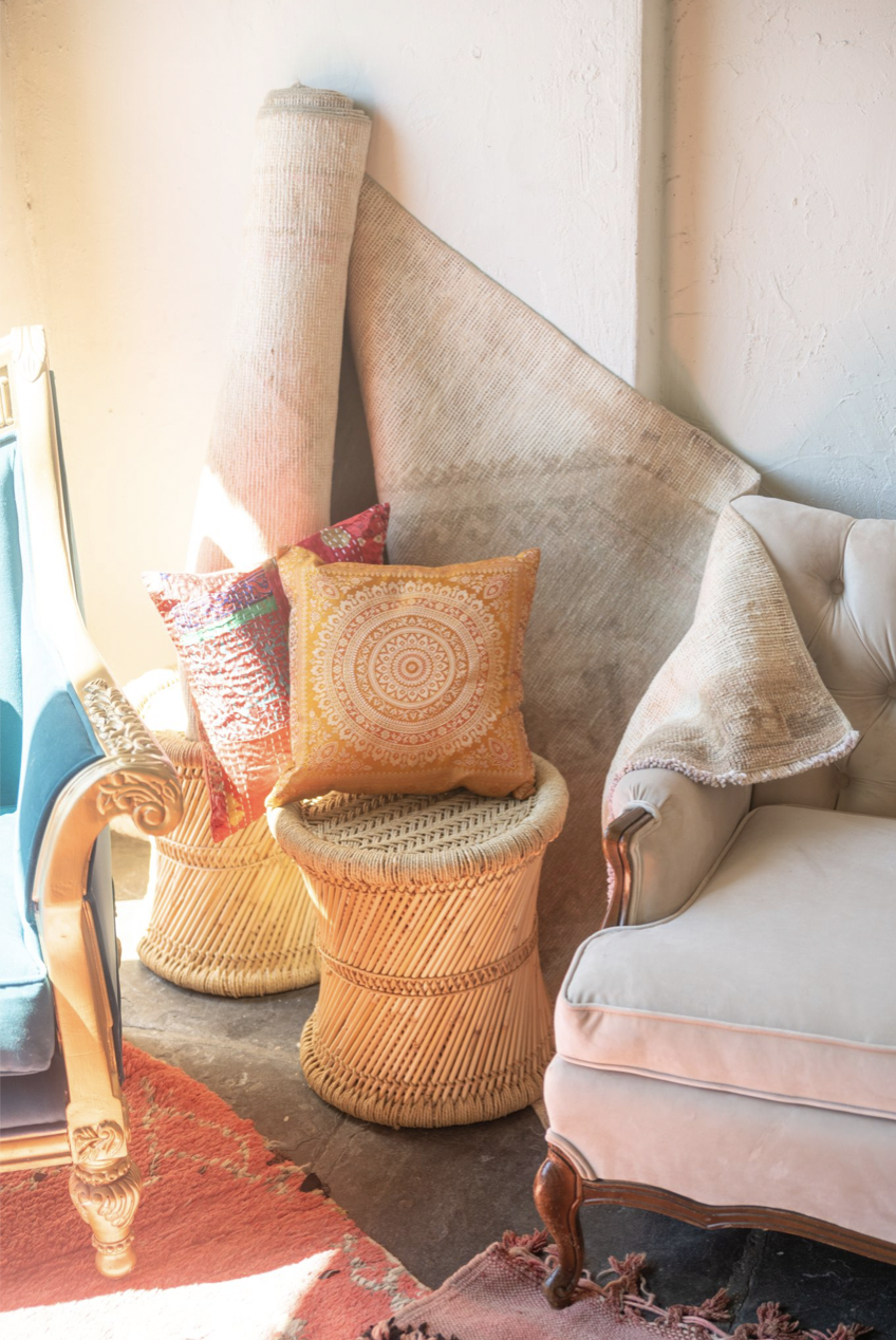 A cozy sitting area with a vintage armchair, a rattan side table, textured cushions, a rolled-up rug, and a large textured fabric piece leaning against the wall, illuminated by warm natural light.