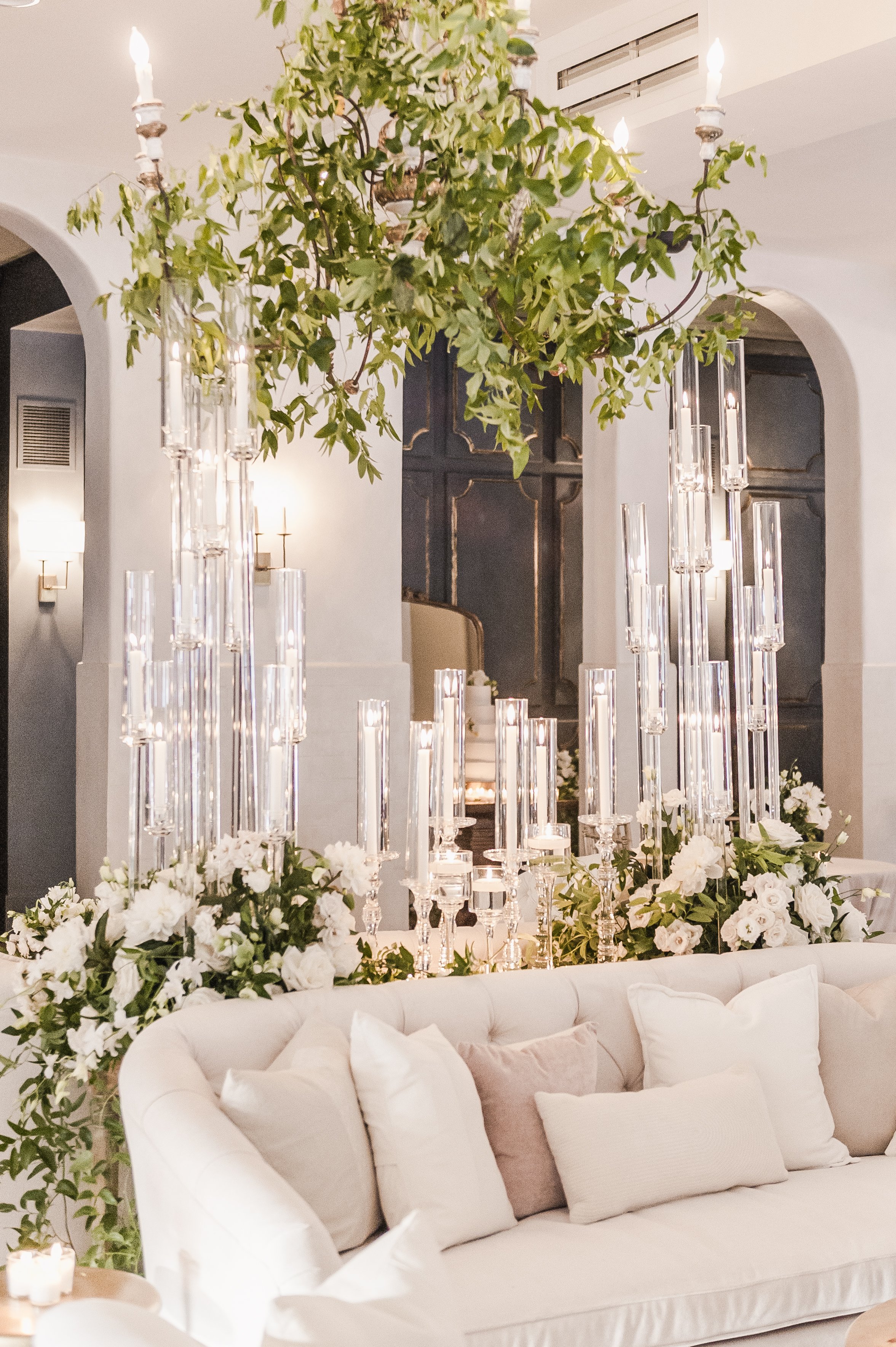 An elegant indoor space decorated with romantic floral arrangements, tall glass candle holders, and a cozy white sofa with cushions, suitable for a special event or wedding reception.