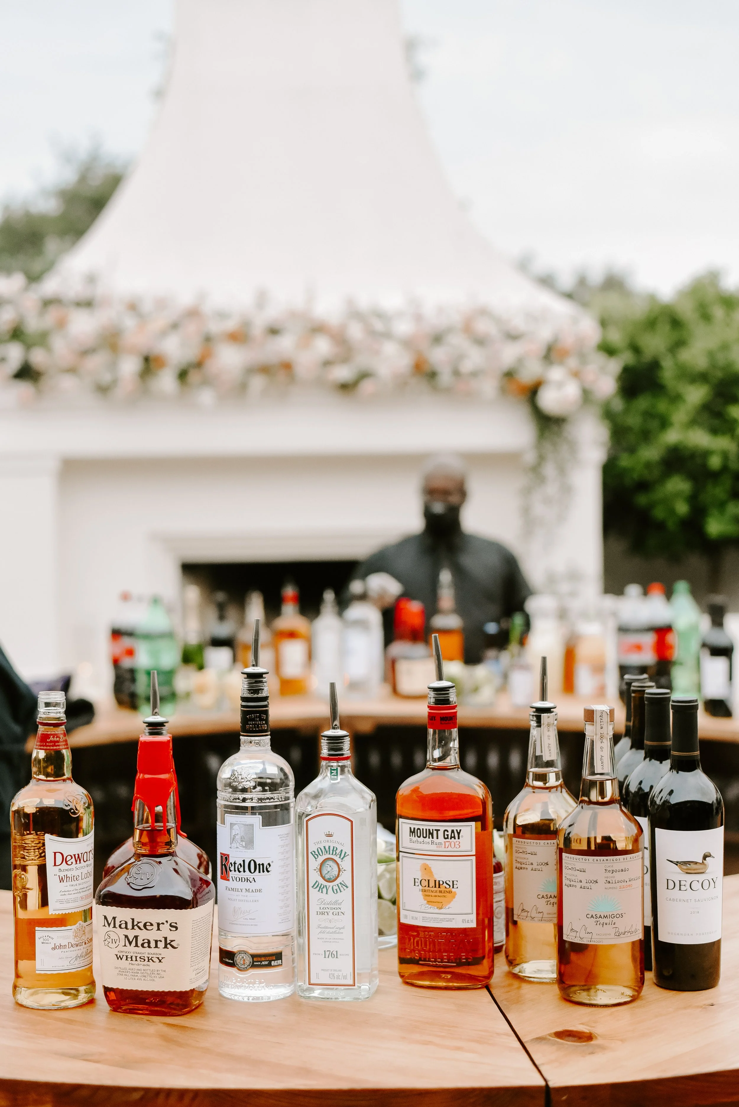 A variety of liquor bottles arranged on a wooden table with blurred background of an outdoor setting, possibly a bar or party.