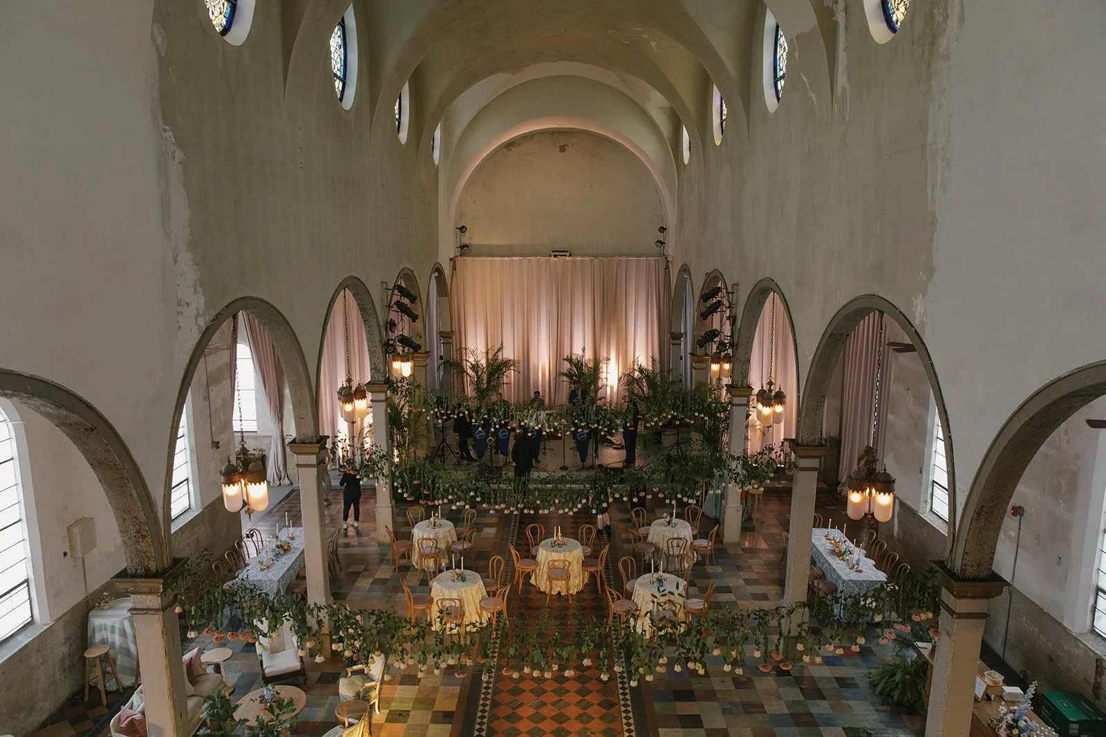Elegant event space with high arched ceilings, tall narrow windows, round tables with yellow tablecloths, and a stage with draped curtains and floral arrangements.