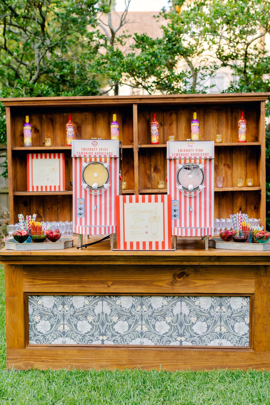 A lemonade stand featuring two striped lemonade dispensers labeled 'Smirnoff Spicy Tamarind Hurricane' and 'Smirnoff Pink Lemonade Hurricane,' with colorful straws, small bottles on top, and bowls of snacks, set against a wooden backdrop with greener