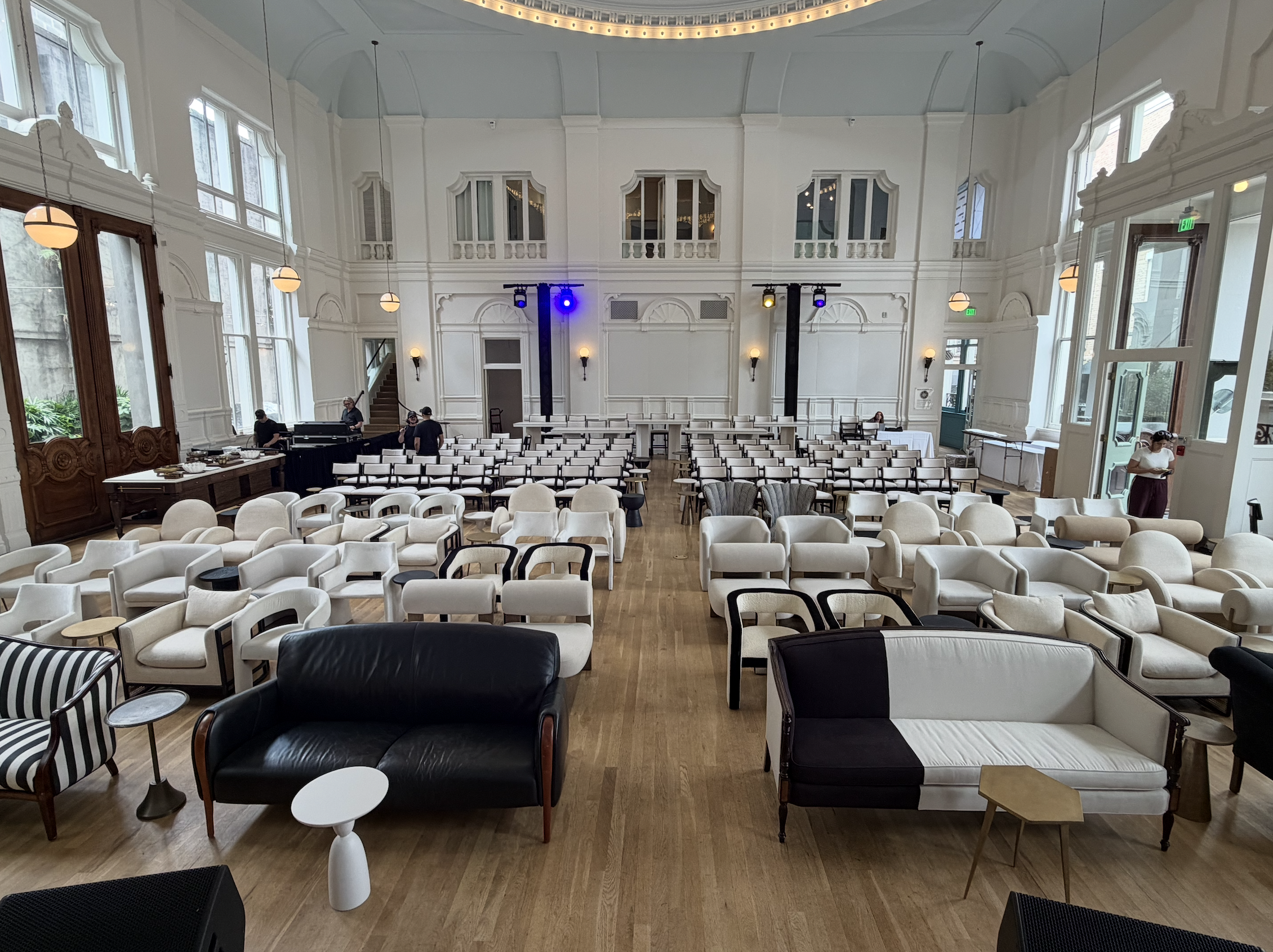 Elegant event space with rows of white and beige chairs, black sofas, and various tables arranged in front of a stage with blue and yellow lighting. Large windows with natural light, ornate white walls, and a ceiling with strip lighting contribute to
