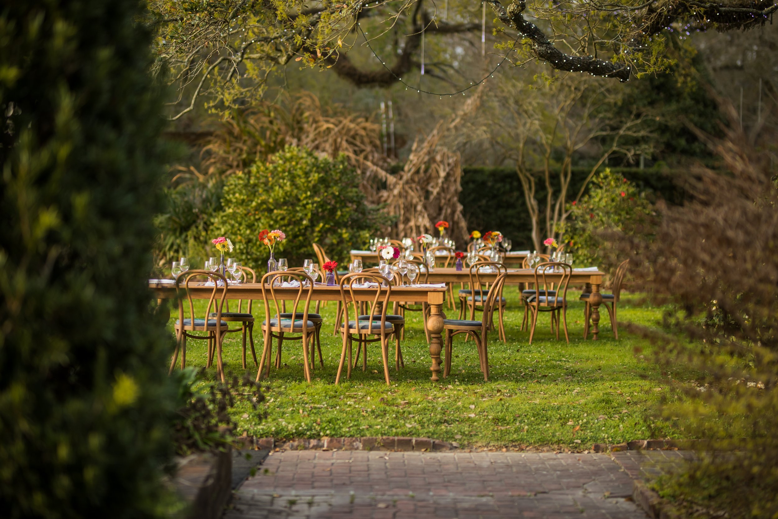 Outdoor wedding or event setup with tables and chairs decorated with colorful flowers, viewed through a natural frame of bushes and trees.