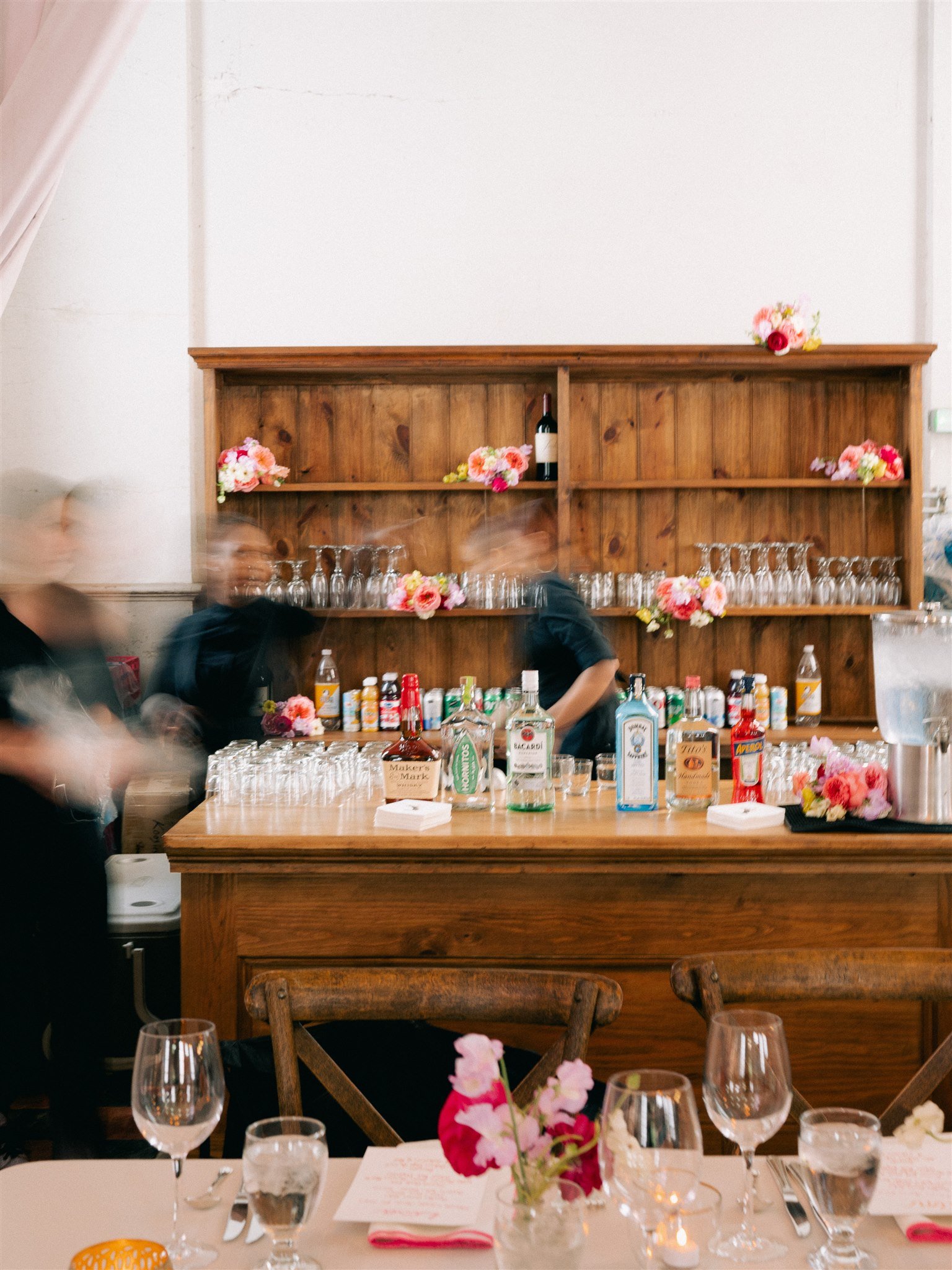 A decorated table set for a celebration with multiple glasses, wine bottles, floral centerpieces, and menu cards. In the background, a wooden bar with floral arrangements and bottles of alcohol is visible.