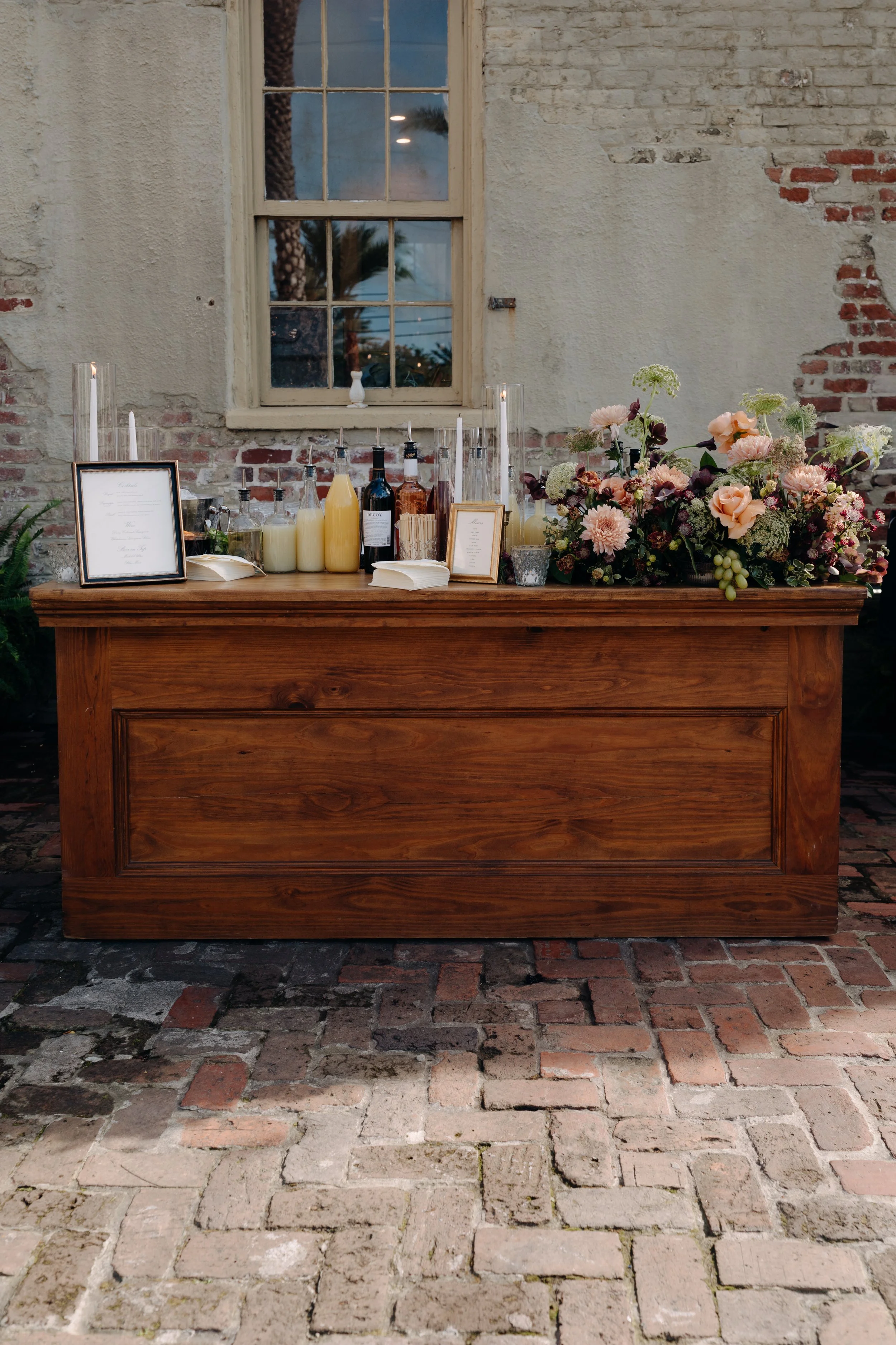 A rustic wooden table with floral arrangements, framed menus, bottles of drinks, and candles, set against a distressed brick and plaster wall with a window showing palm trees outside.