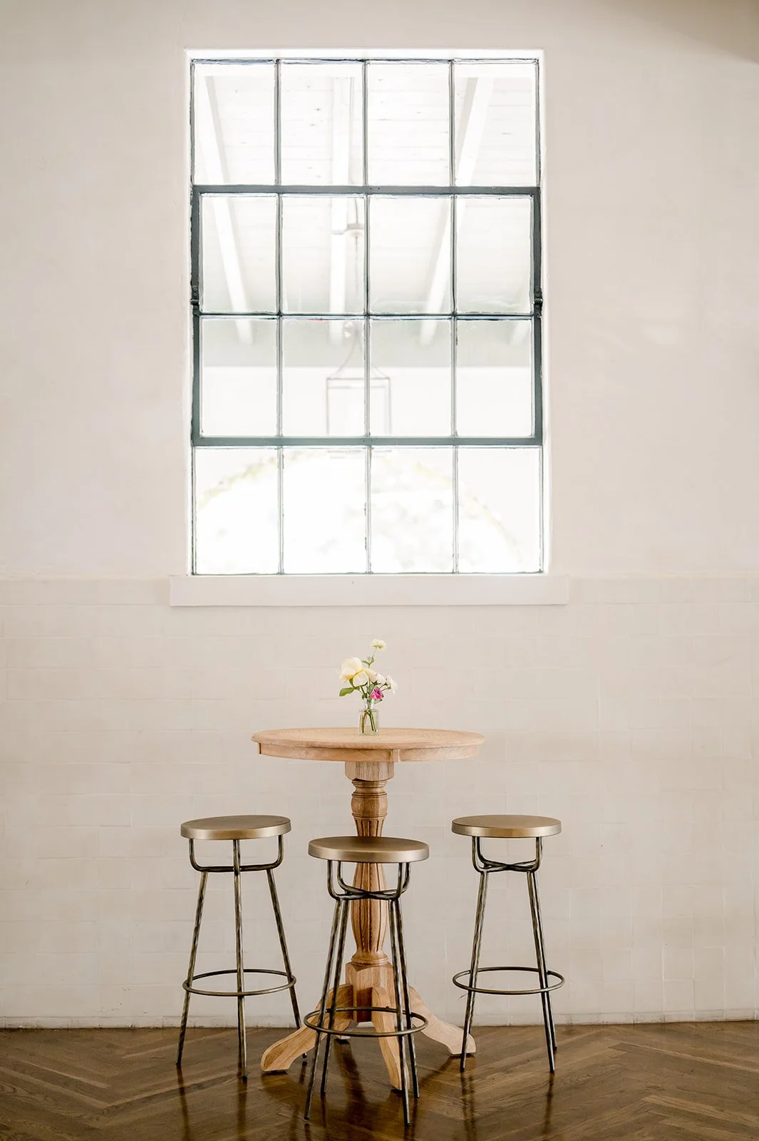 A wooden table with three barstools and a small vase of flowers, set against a white wall with a large window overhead that lets in natural light.