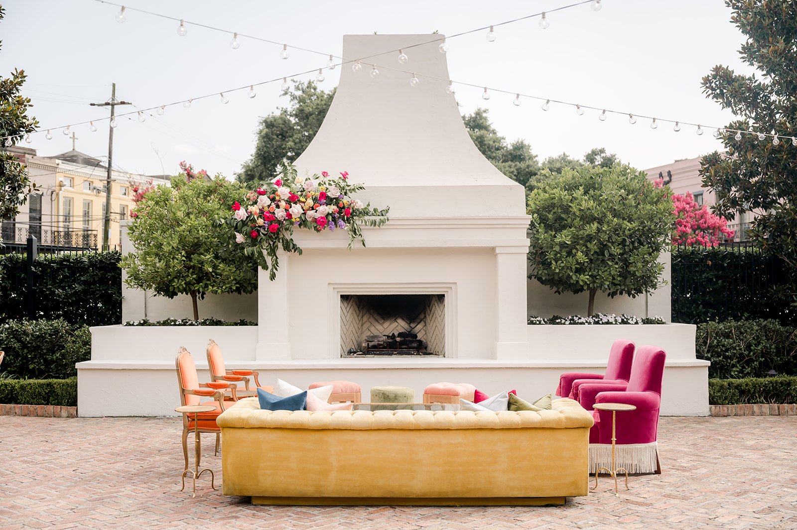 Outdoor seating area with a white fireplace, pink and orange chairs, pink armchairs, colorful pillows, and string lights above. Flowering bushes and trees surround the space.