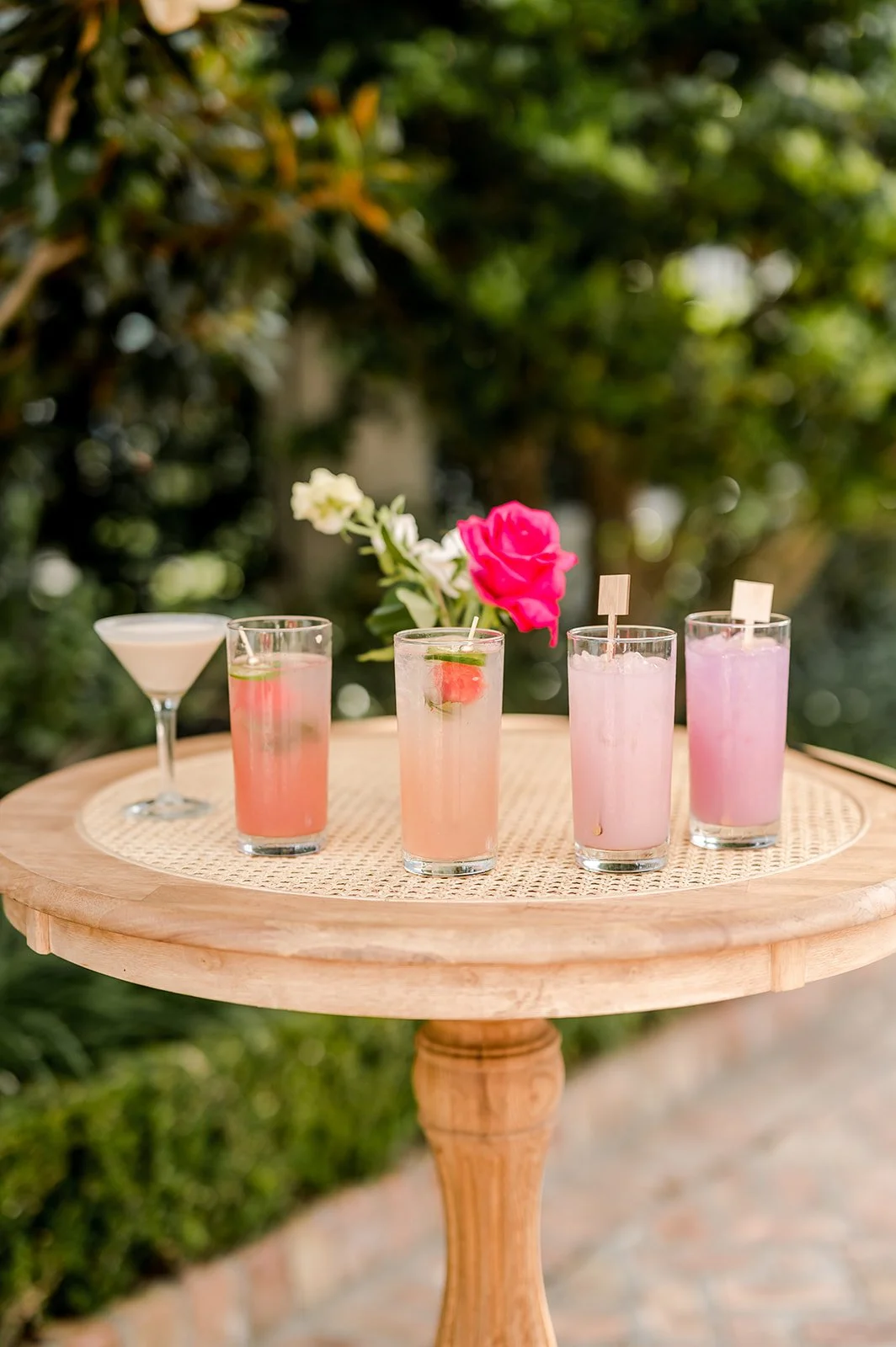 Five colorful cocktails arranged on a round wooden table outdoors with greenery in the background, including a pink rose and white flowers.