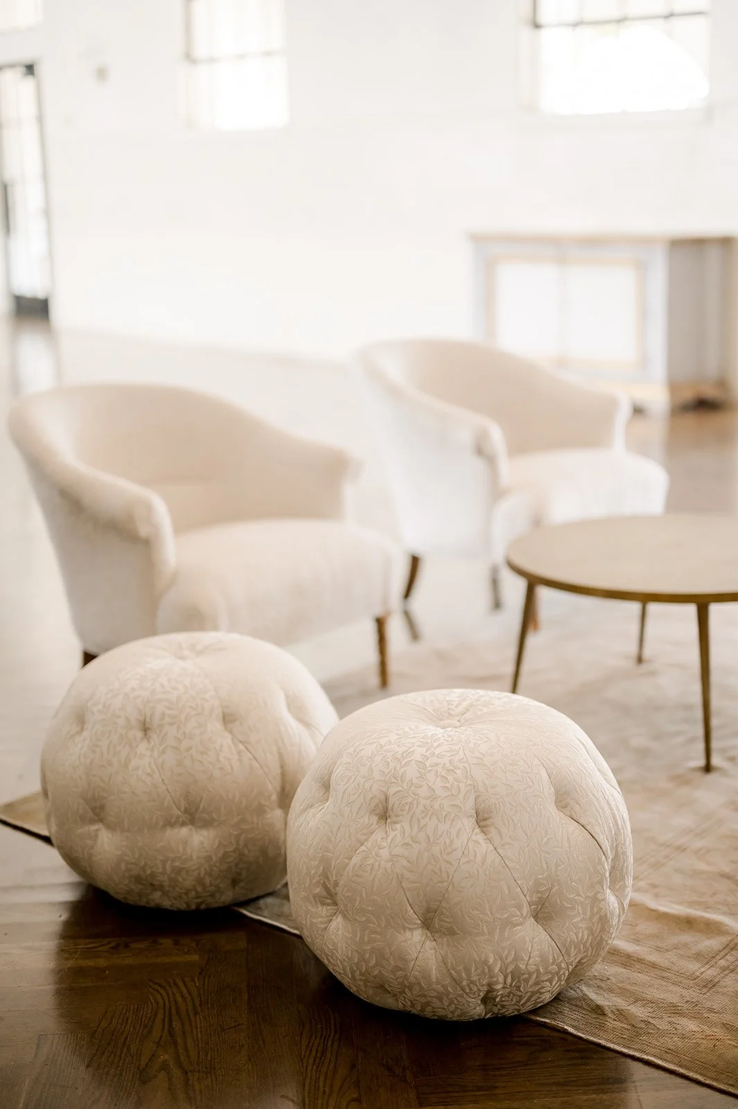 Living room with two white armchairs, two round beige tufted ottomans, a wooden coffee table, and a white sideboard against the wall.