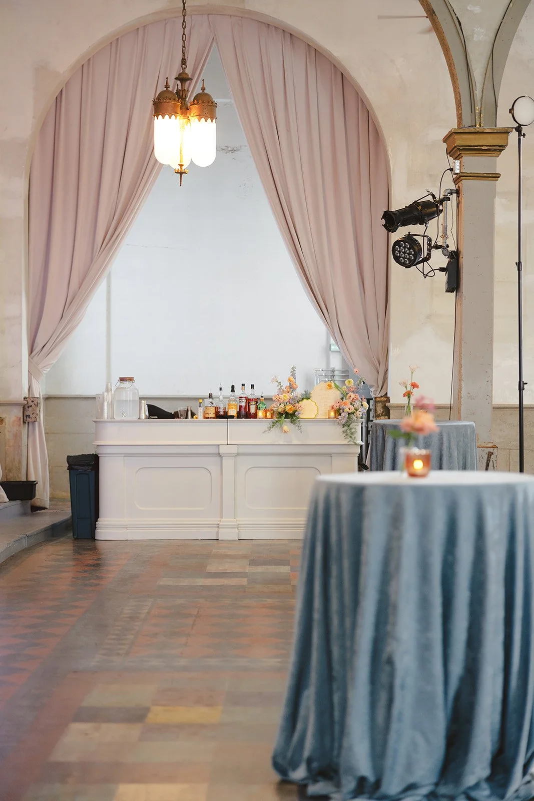 Event space with a bar area behind a white counter, decorated with flowers and drinks. Elegant pink curtains frame an arched opening, and tables with blue velvet tablecloths and candle centerpieces are in the foreground. Stage lighting and a vintage 