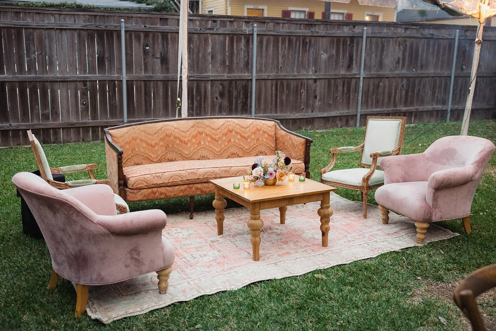 Outdoor seating arrangement on a backyard lawn with vintage-style furniture including a peach sofa, pink armchairs, and green chairs surrounding a wooden table with candles and flowers, enclosed by a wooden fence.
