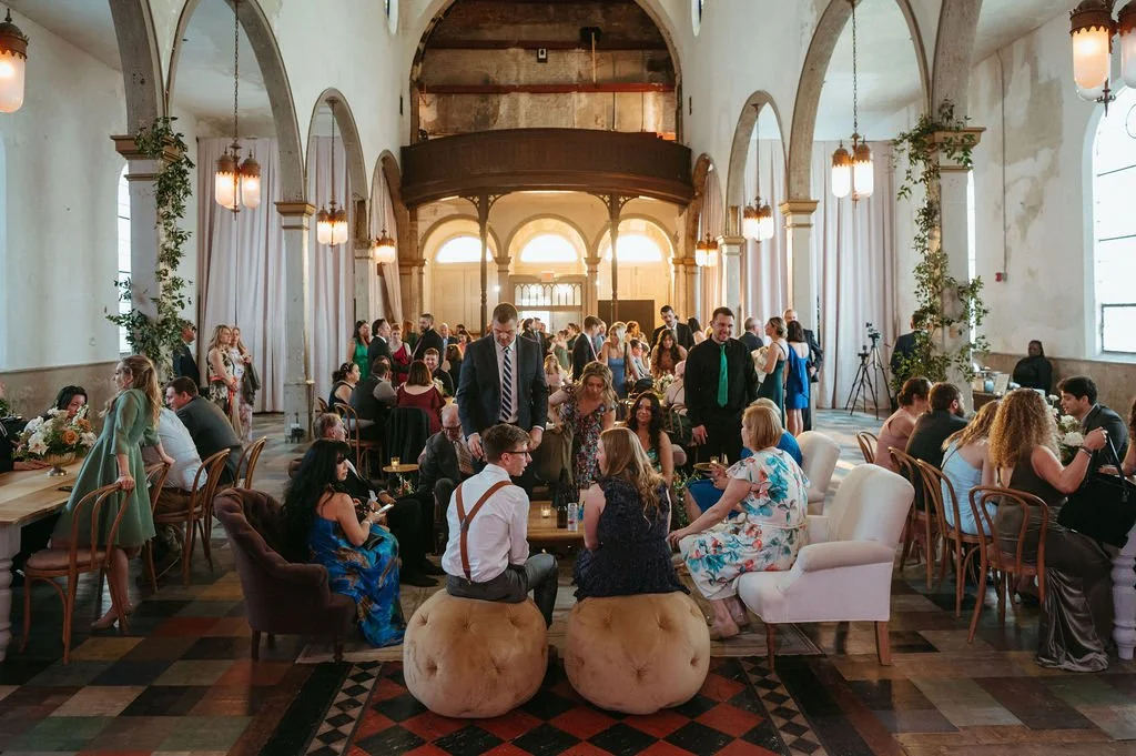 A large indoor social gathering or party with many people seated and standing in a spacious, vintage-styled hall with arched windows and hanging lights.