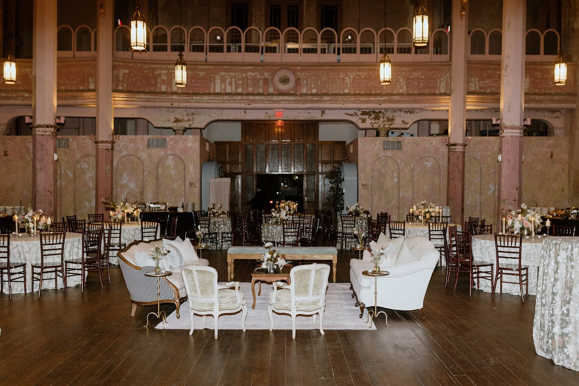 Elegant event space with round tables covered in white lace tablecloths, decorated with candle centerpieces, surrounded by wooden chairs. The room has a high, ornate ceiling with hanging lantern-style lights, exposed plaster walls with peeling paint,