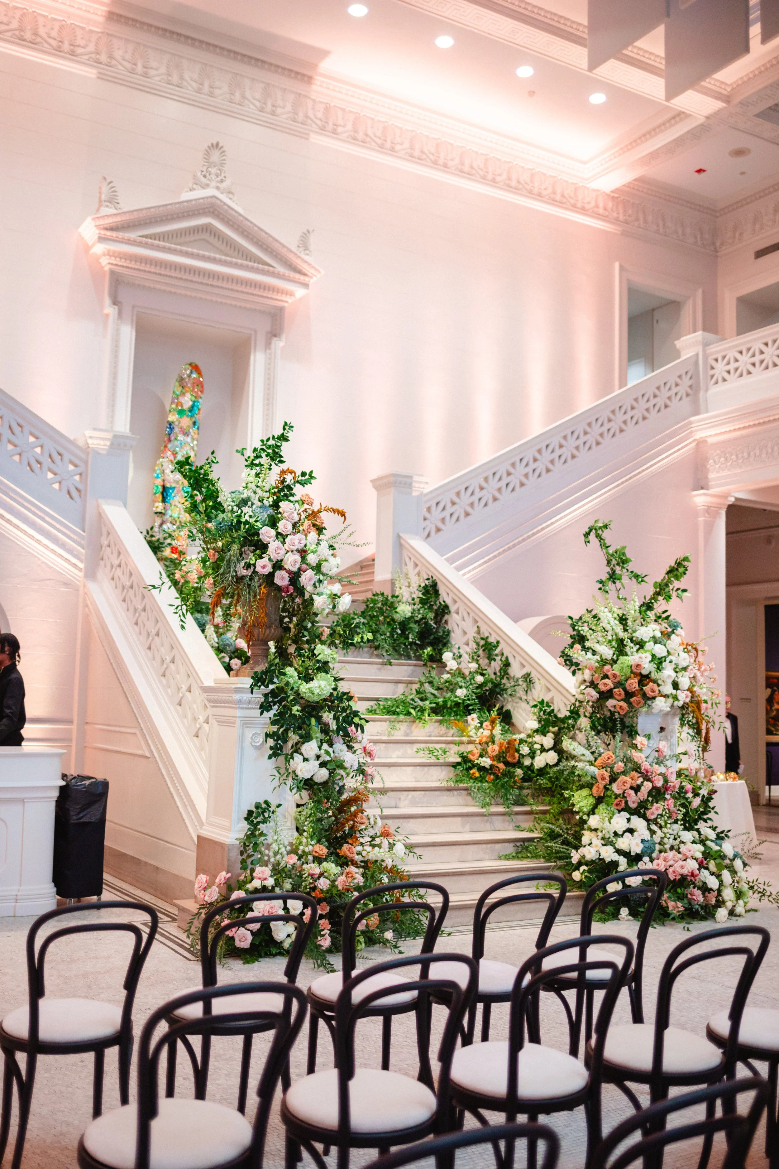 Elegant wedding venue with a grand staircase decorated with colorful floral arrangements, and rows of black chairs with white cushions arranged for guests.