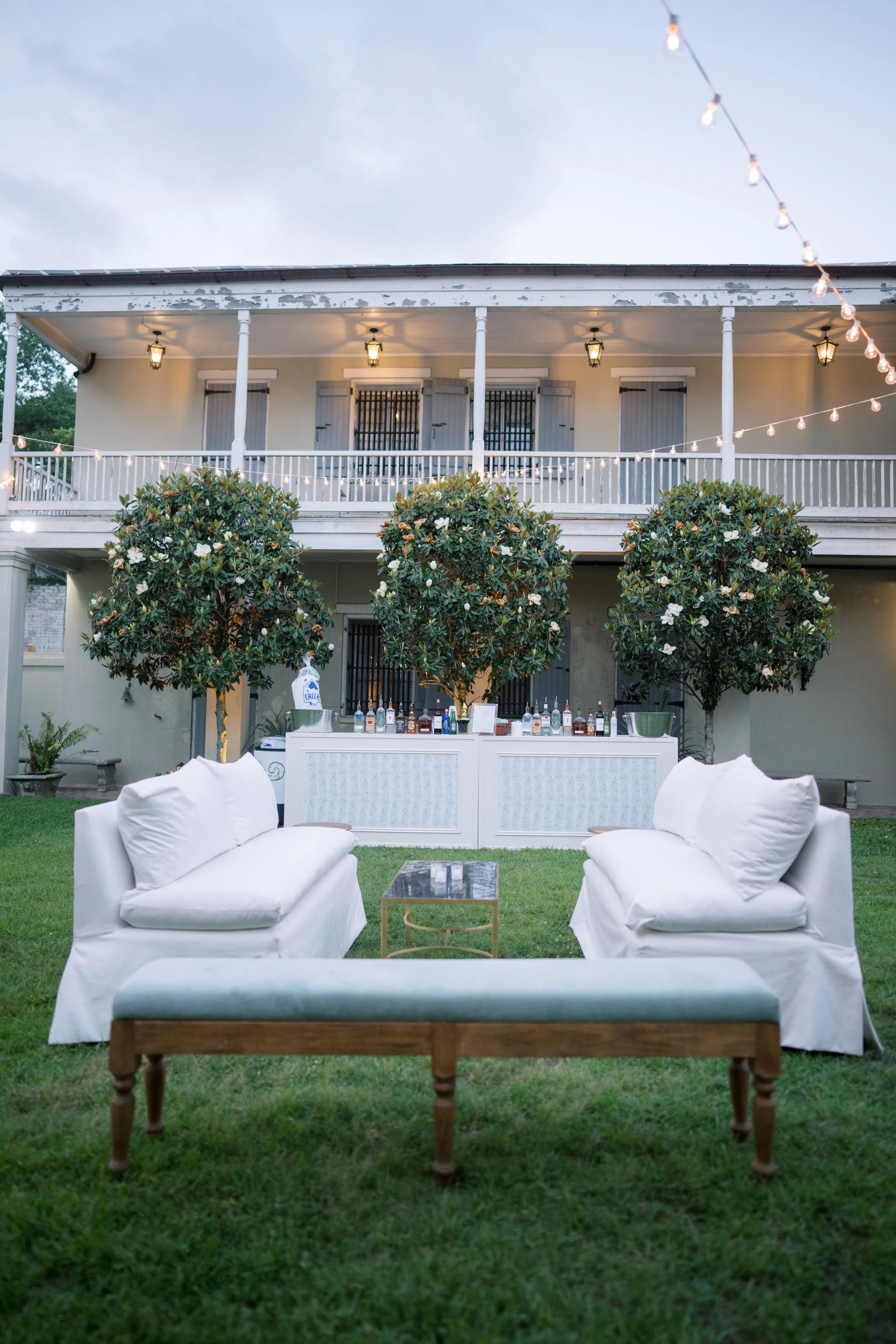 Outdoor backyard setup with white couches, a small table, two trees, a serving bar with drinks, and string lights hanging between the trees, with a large house in the background.