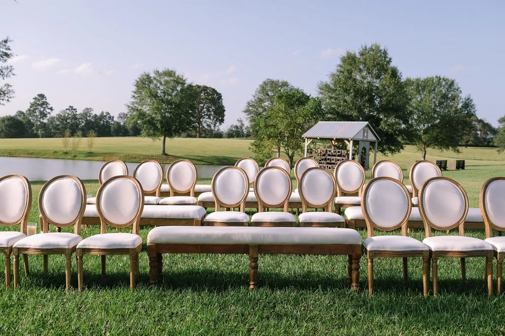Rows of white cushioned chairs arranged outdoors on a lush green lawn, facing a small pond and a backdrop of trees, with a small wooden structure in the distance, under a clear blue sky.