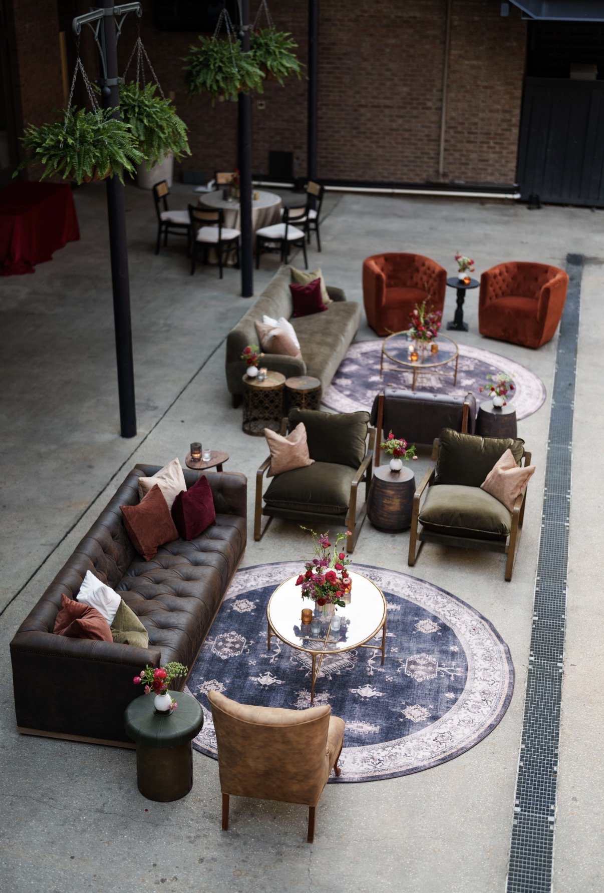 Indoor event space with vintage-style furniture including sofas, armchairs, and round tables decorated with flowers, candles, and decorative pillows. The space has a concrete floor, brick walls, and hanging green plants.