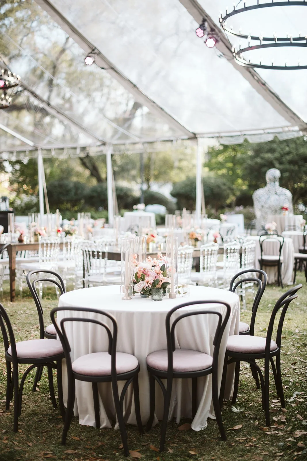 Decorated outdoor event space with round tables draped in white tablecloths, pink floral centerpieces, and surrounded by black chairs, set under a transparent canopy with hanging lights and trees in the background.