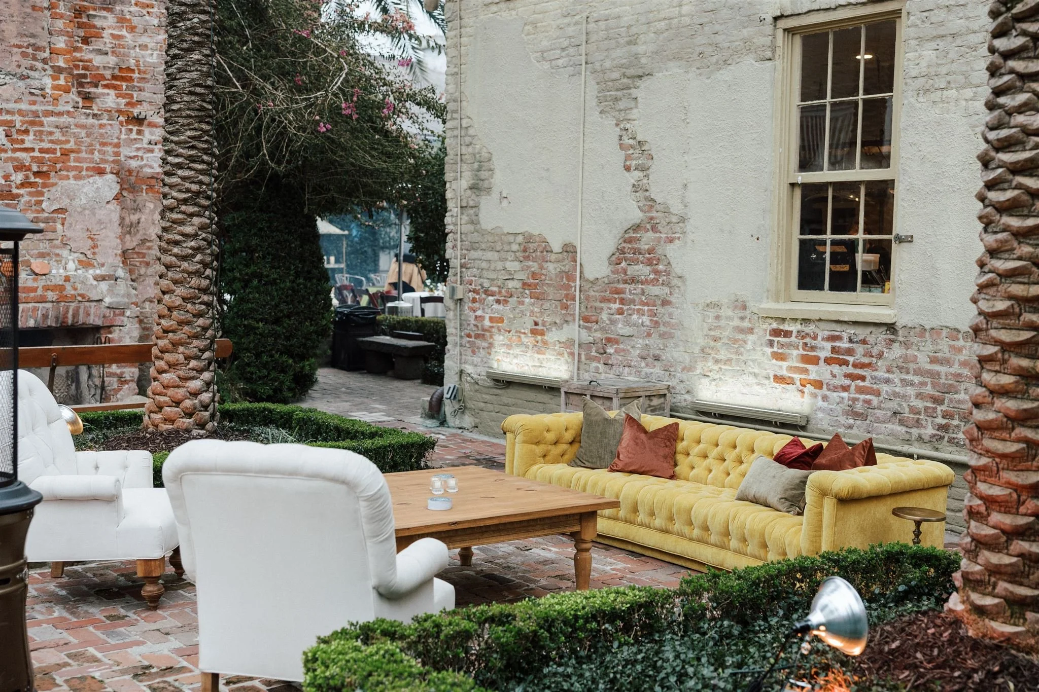 A cozy outdoor patio with vintage-style furniture, including a white chair and a yellow tufted sofa with pillows, surrounded by brick walls, greenery, and trees.