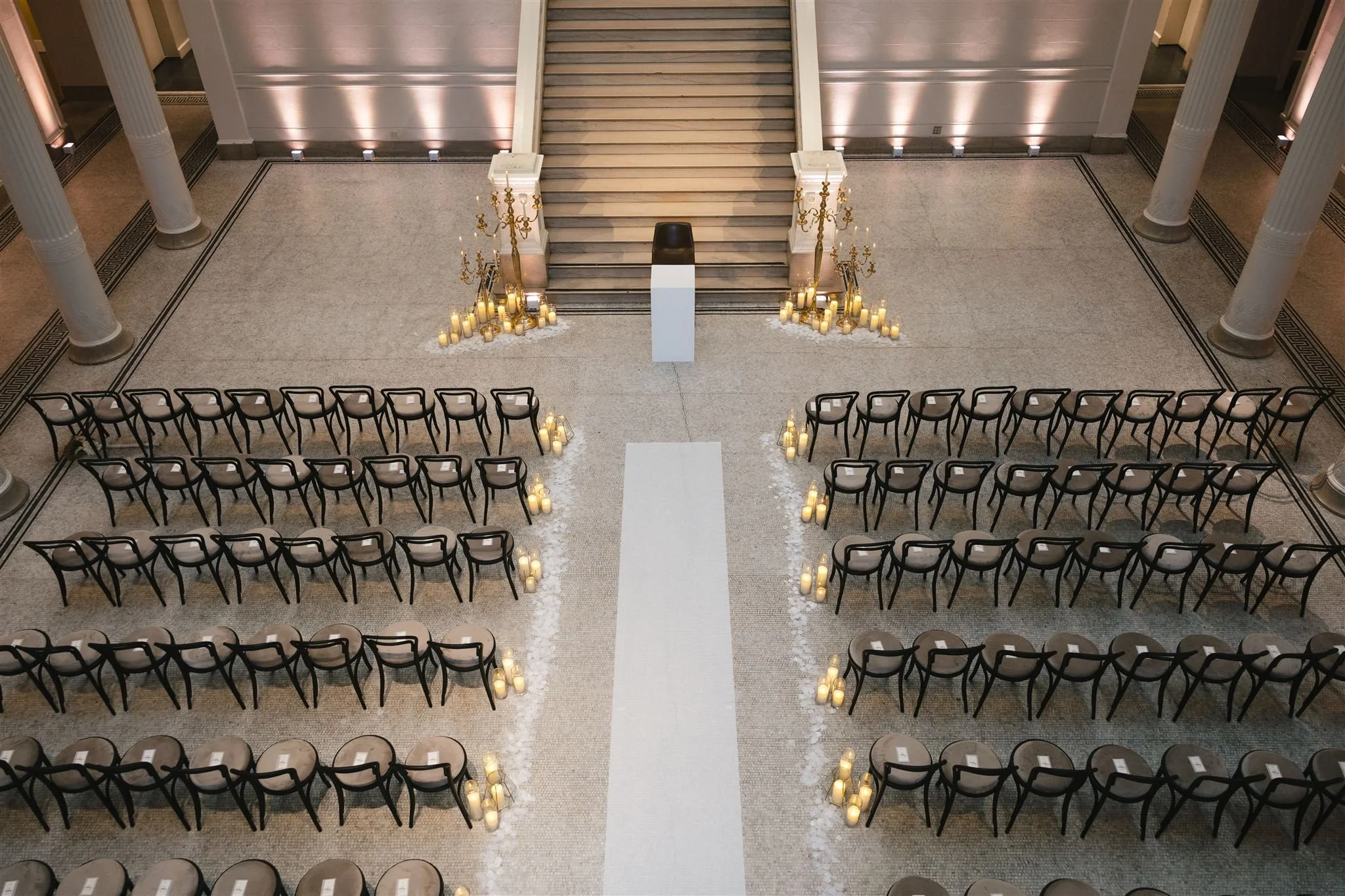 Interior of a grand hall with rows of chairs arranged for a formal event, featuring candles and chandeliers, a staircase at the center, and decorative columns.
